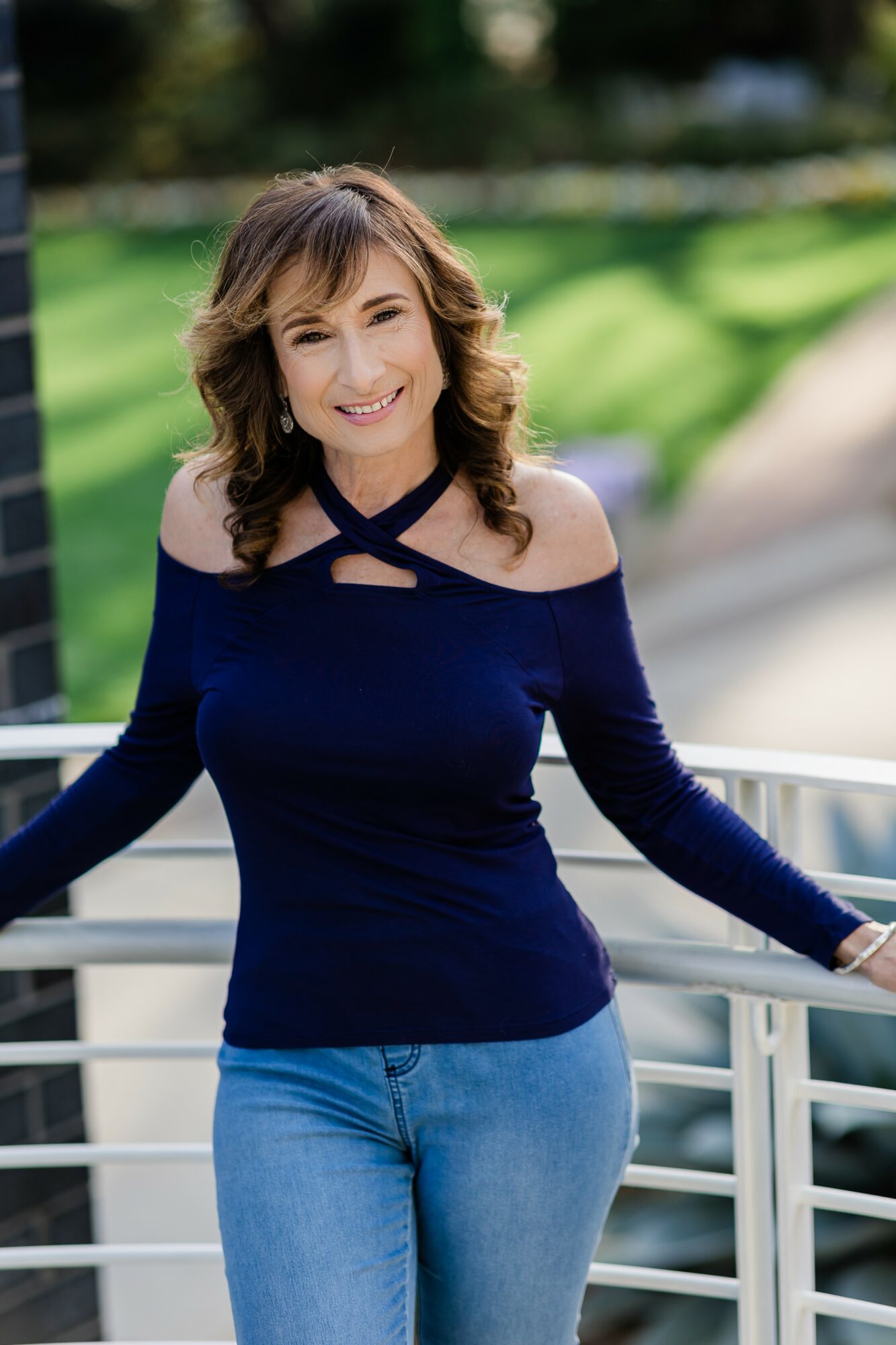 Woman with shoulder-length hair smiling outdoors, wearing a navy blue top and jeans, leaning on a railing.