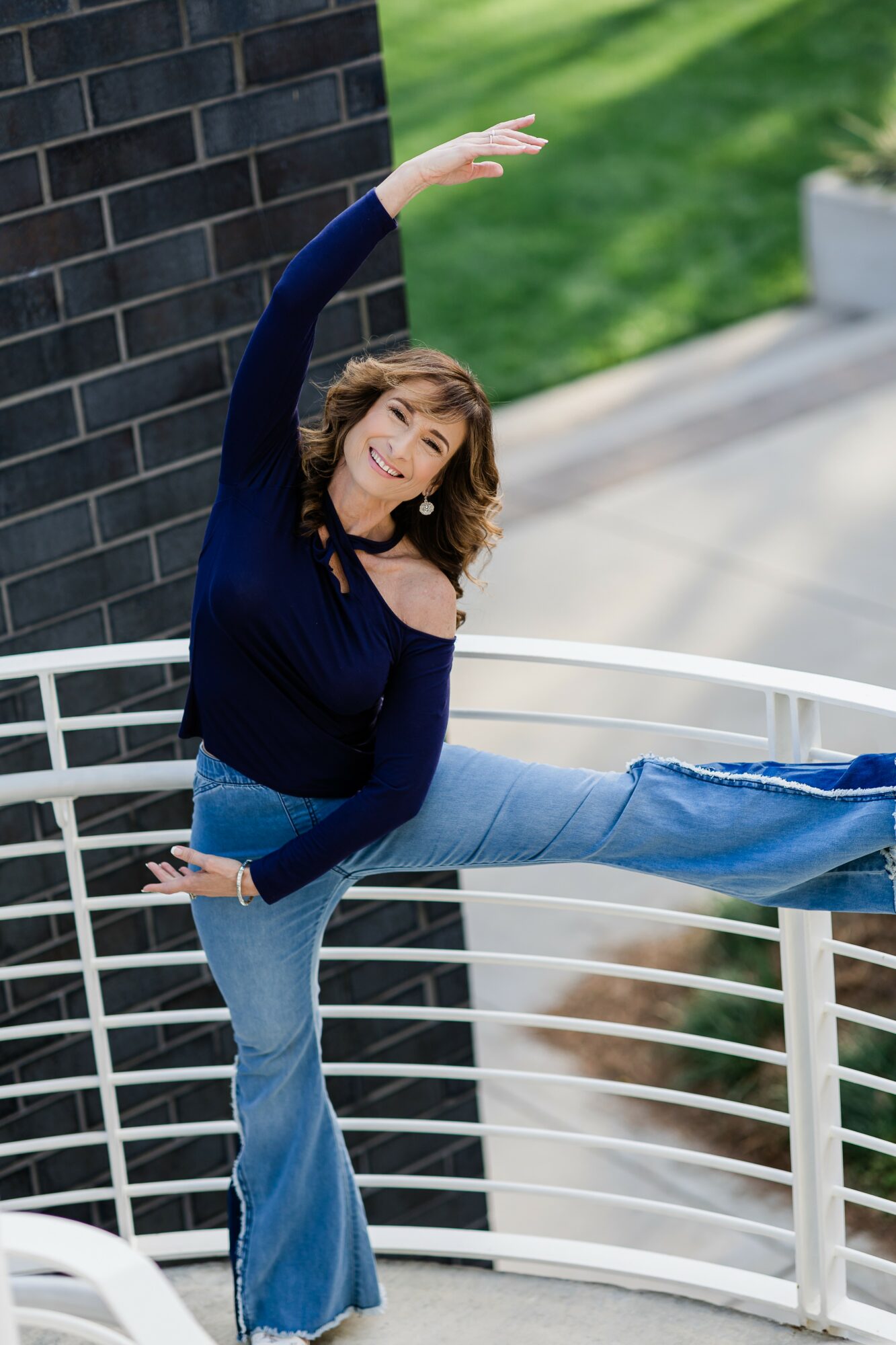 Woman with curly hair stretching her arm and leg outdoors, leaning against a black brick wall, smiling.