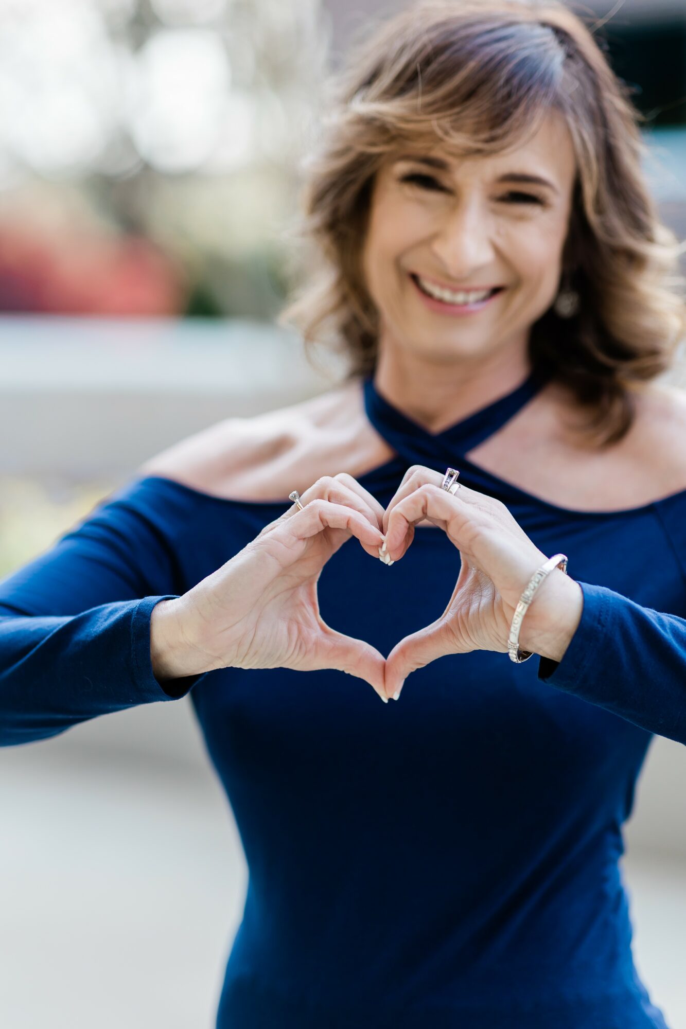 Smiling woman outdoors making a heart shape with her hands, wearing a dark blue top, with blurred background.