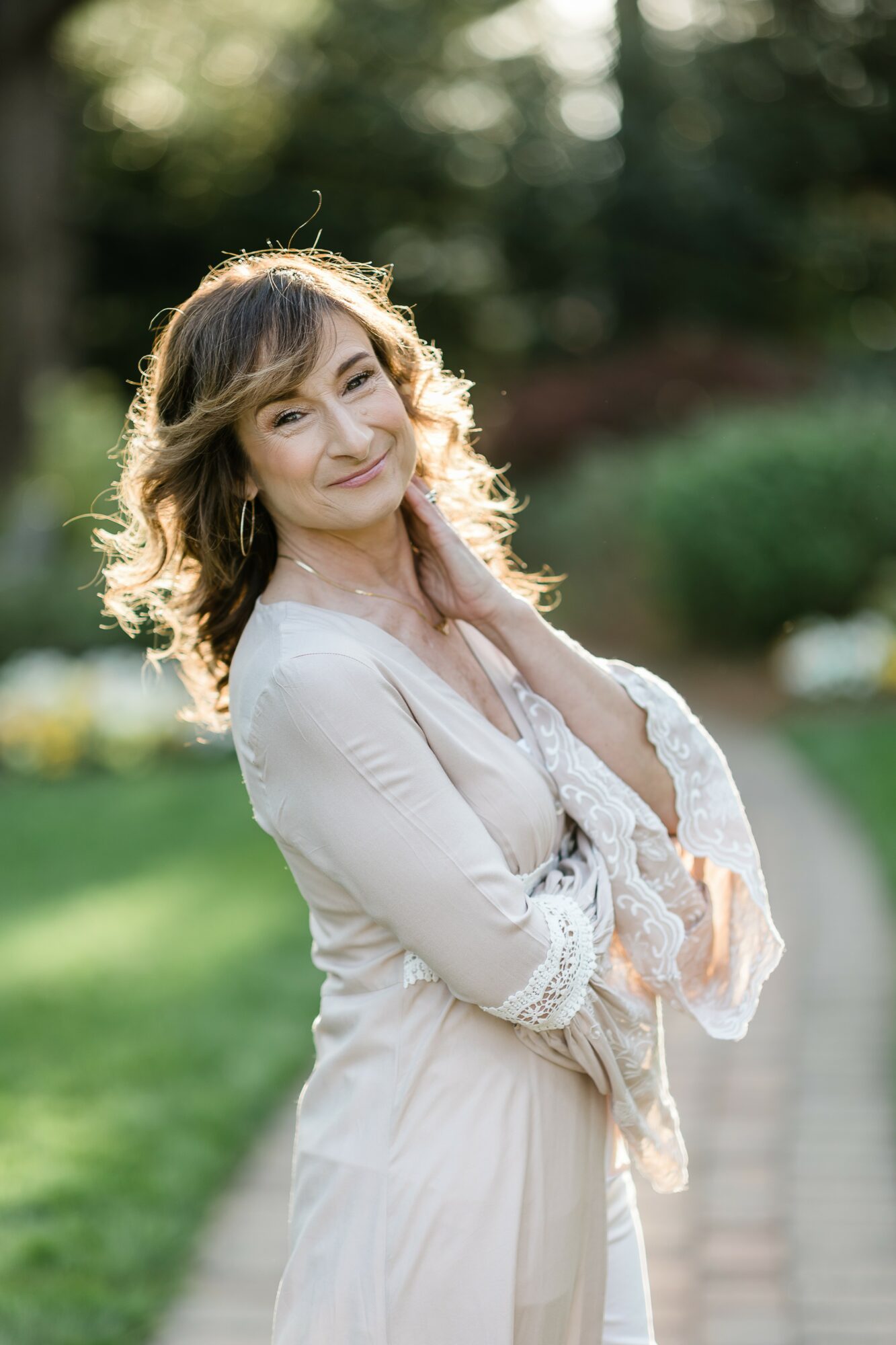 Young woman with wavy hair smiling outdoors, wearing a light-colored dress, standing on a pathway with greenery in background.