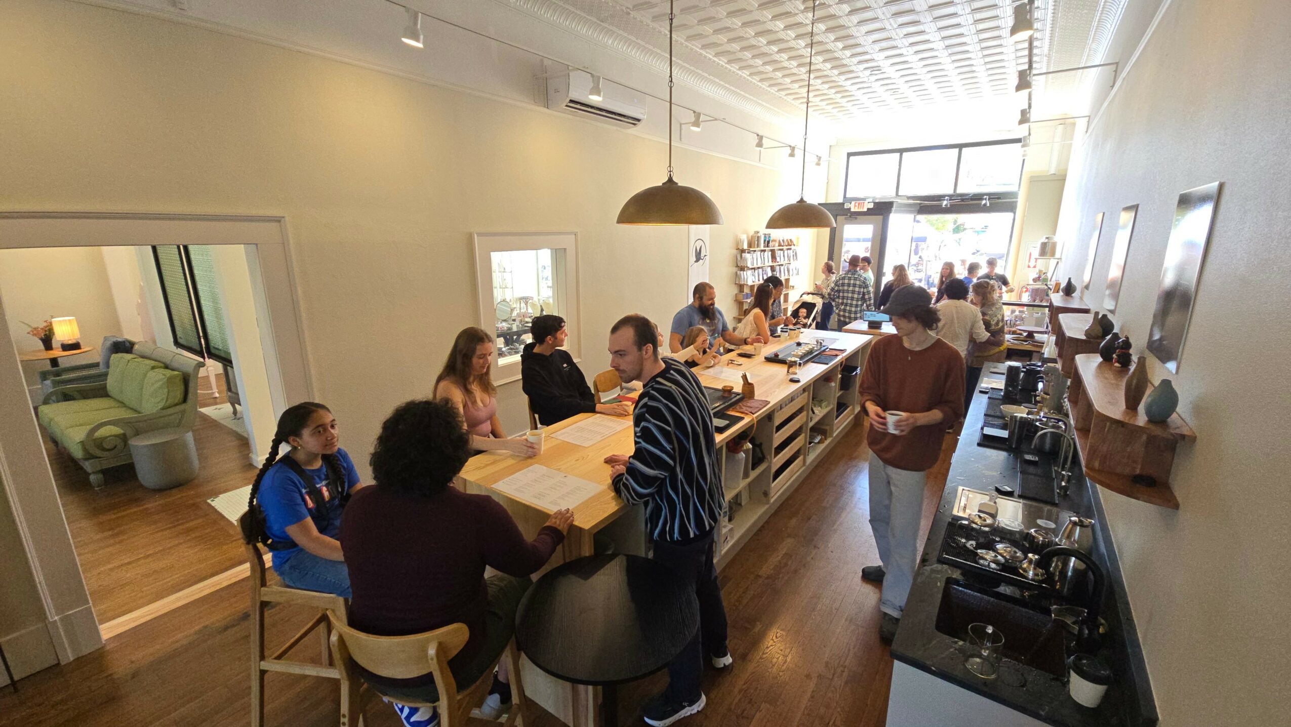 People gathered around a long table in a bright room with large windows, some standing and some sitting, with a kitchen area nearby.