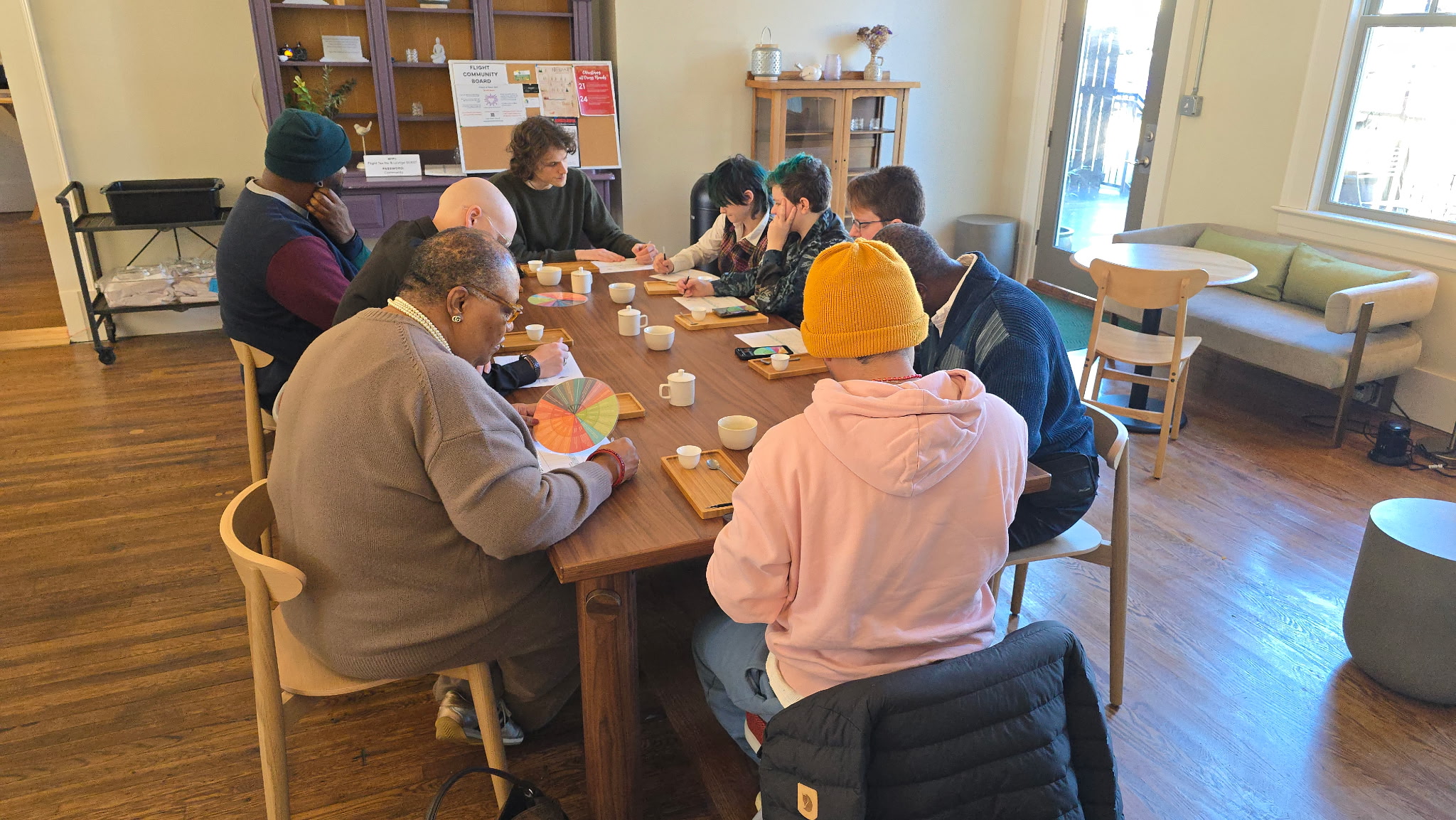 Group of people sitting around a table in a room with wooden floors and shelves, engaged in a discussion or activity.