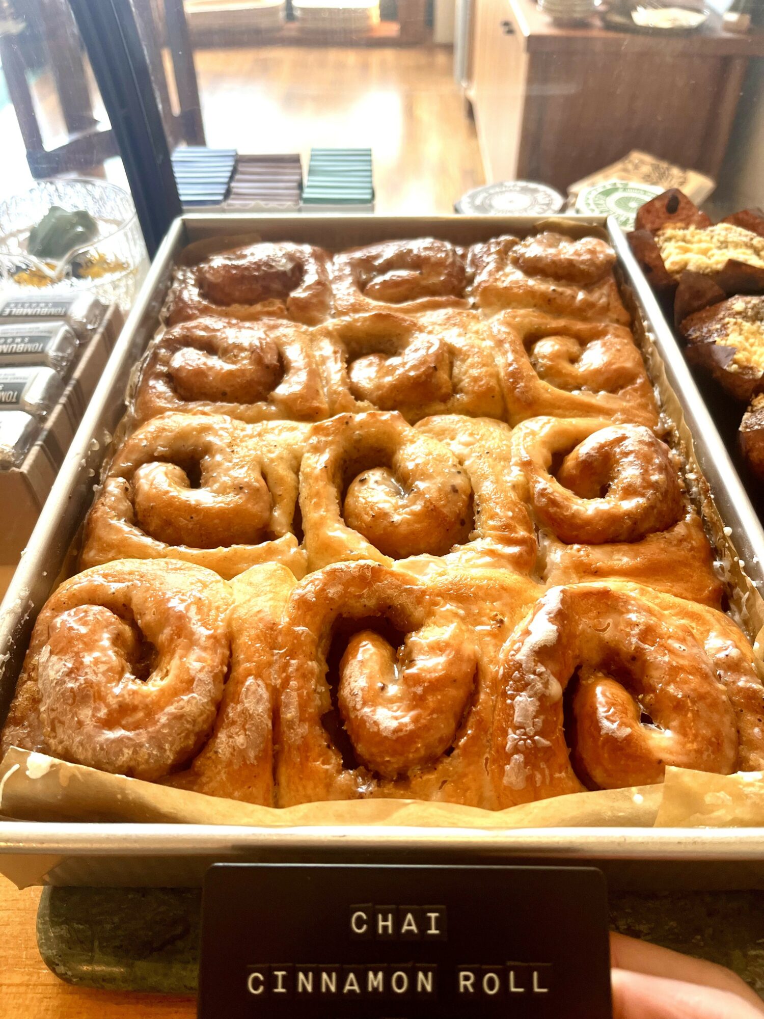 Tray of glazed cinnamon rolls with a person holding a sign that reads 'Chai Cinnamon Roll' in front.