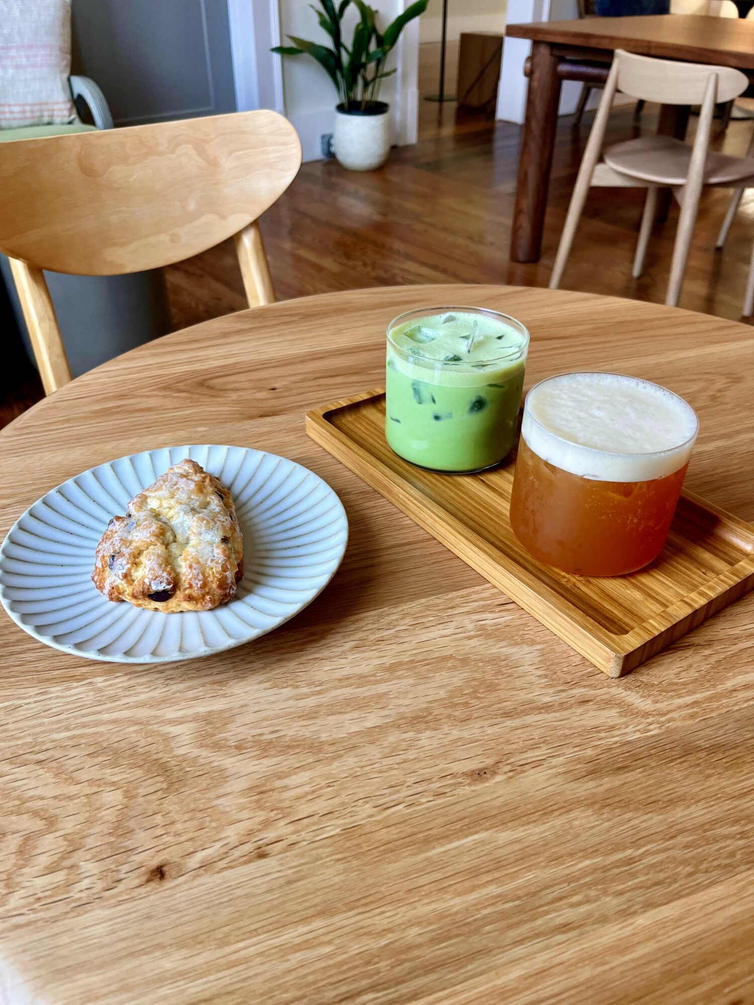 A wooden table with a plate of cake and two drinks on a wooden tray in a cozy room with chairs and a plant.