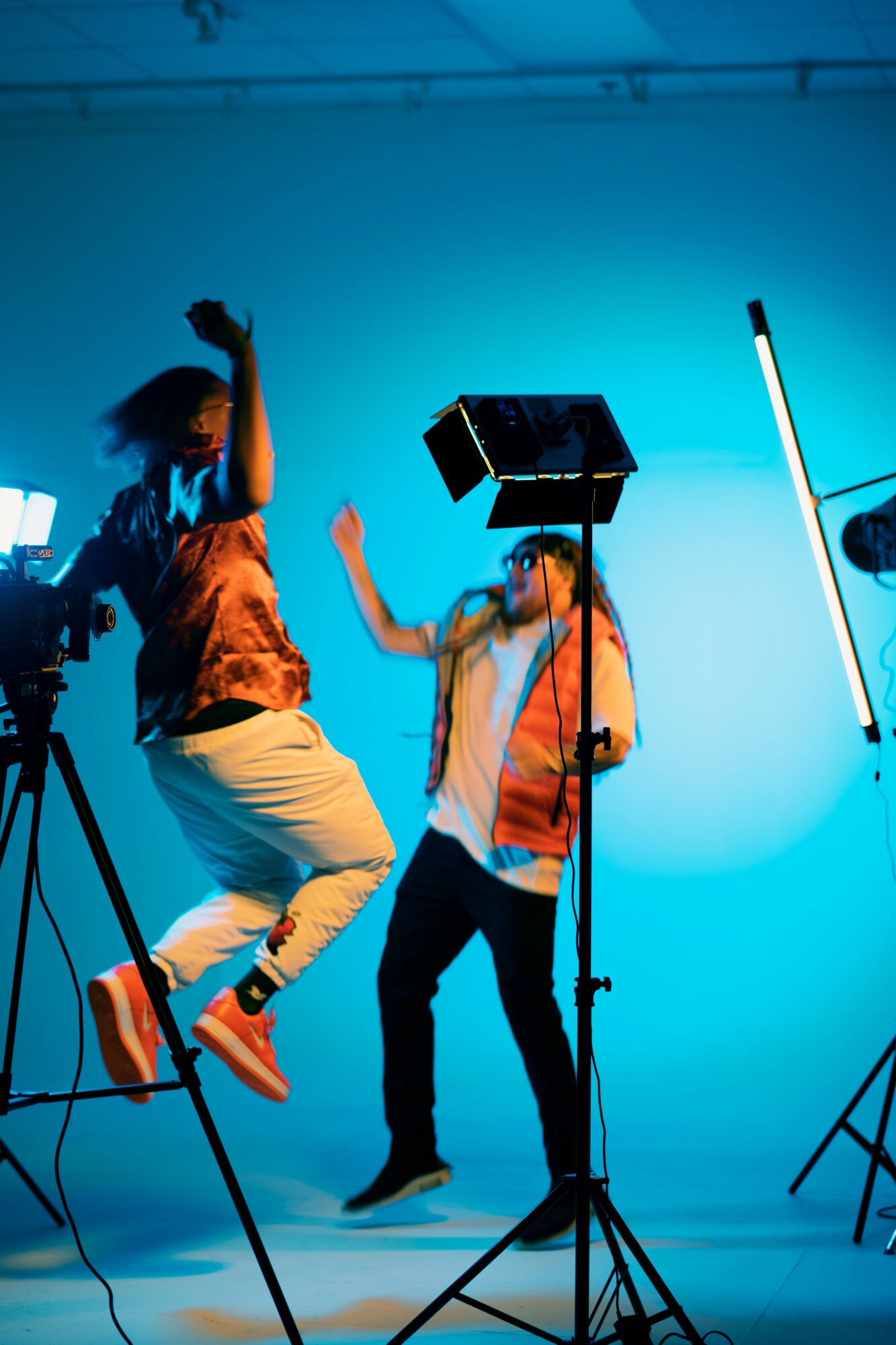Two people dancing in a studio with blue background, one jumping and the other standing near lighting equipment.