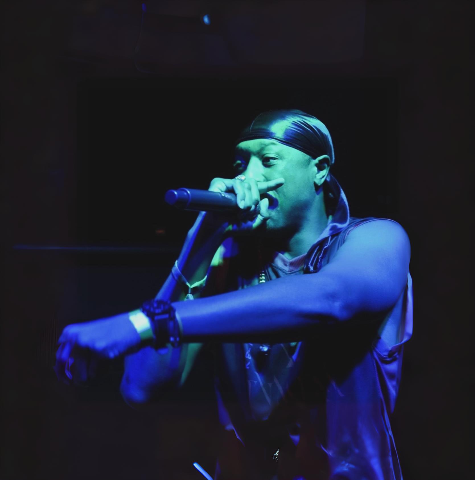 Person singing into a microphone on stage, wearing a headband and sleeveless shirt, illuminated by blue light.