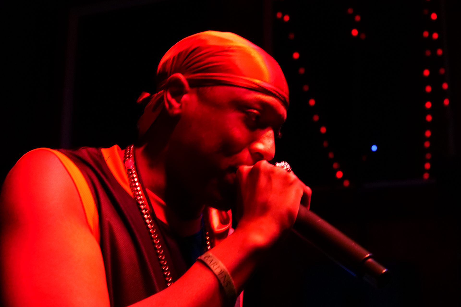 Person wearing a headscarf and jewelry singing into a microphone in a dark setting with red lighting.