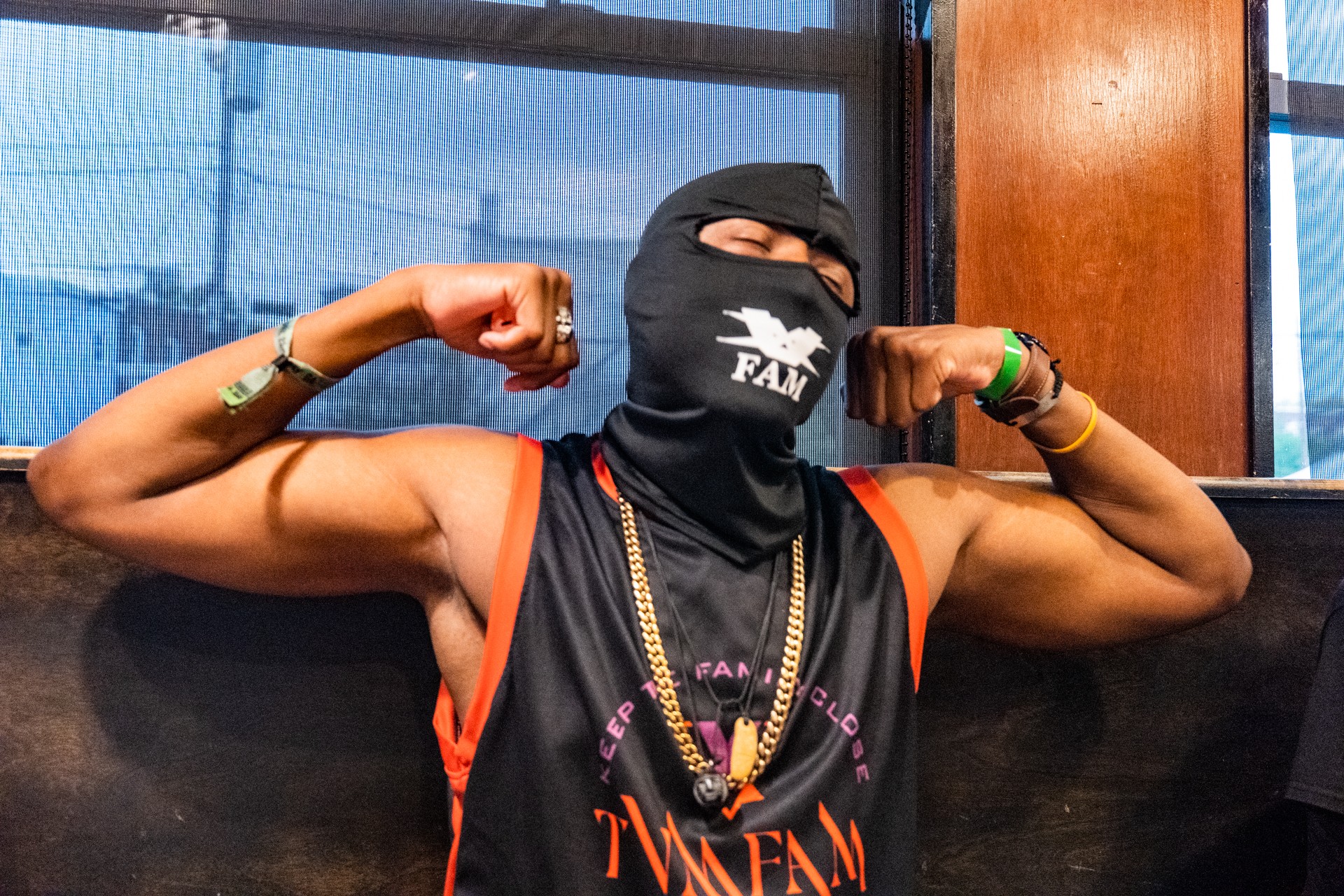 Person wearing a black mask and sleeveless shirt, flexing arms, sitting indoors with windows behind.