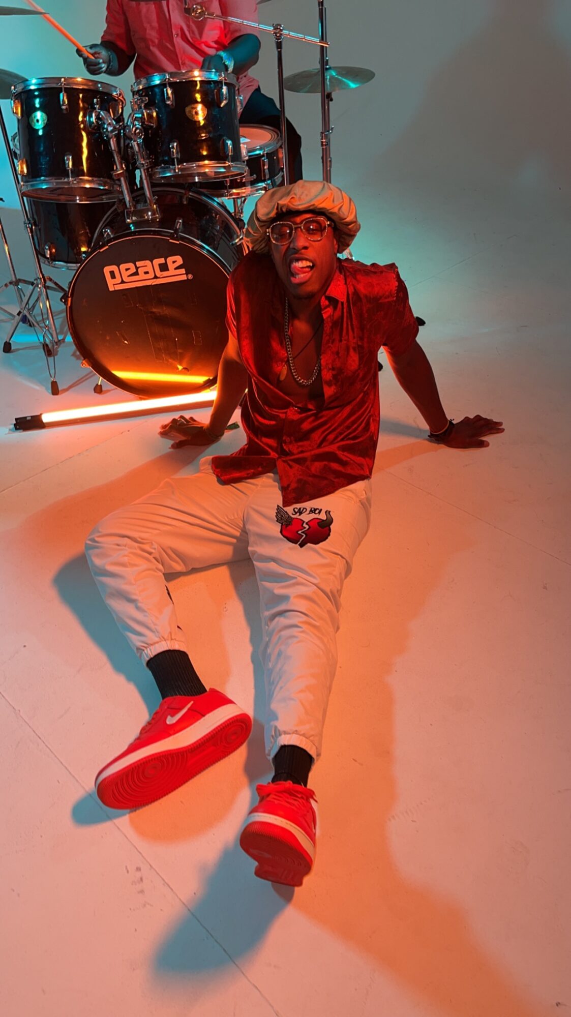 Person sitting on the floor with a drum set in the background, wearing red and beige clothing, red shoes, and sunglasses.