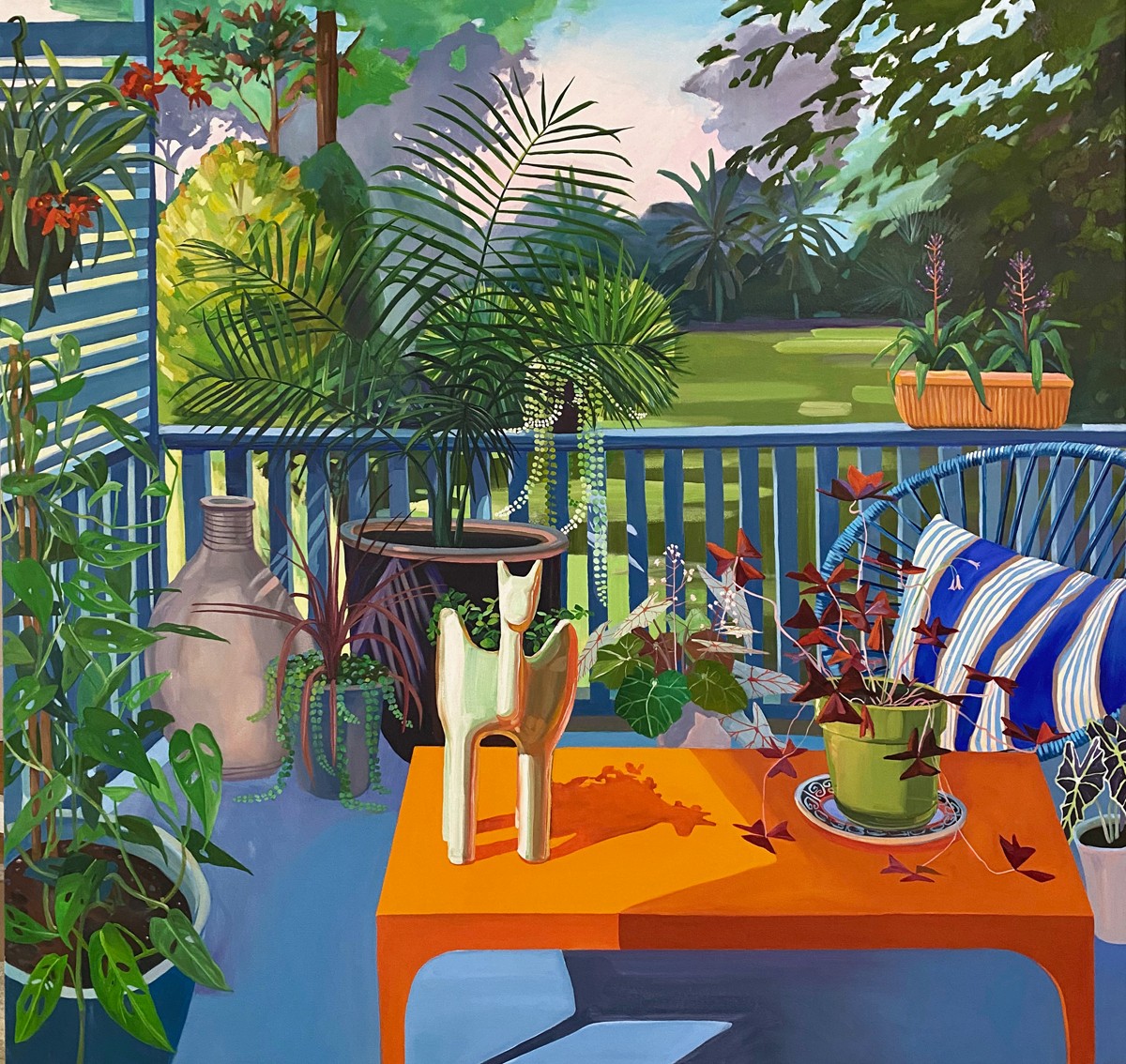 Colorful outdoor table with vases, plants, and striped cushions, overlooking a lush garden with trees and grass.