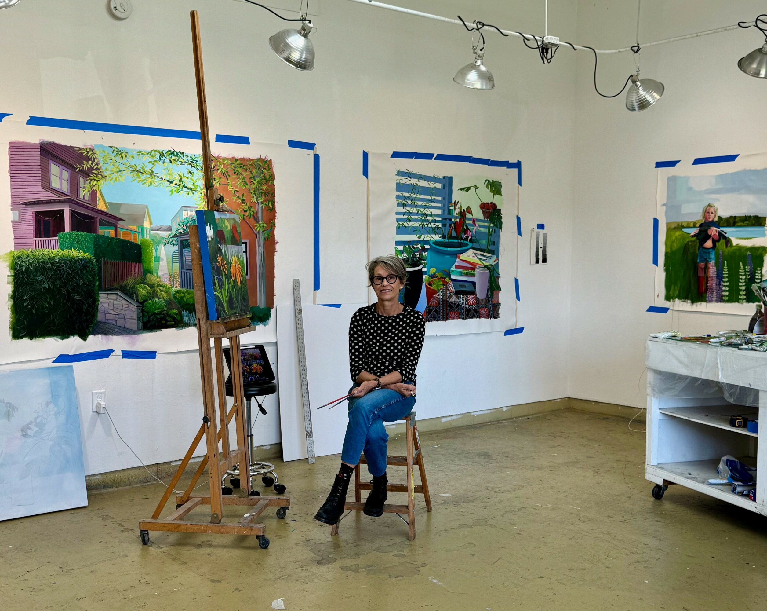 Woman sitting on a stool in an art studio with paintings on the wall behind her.