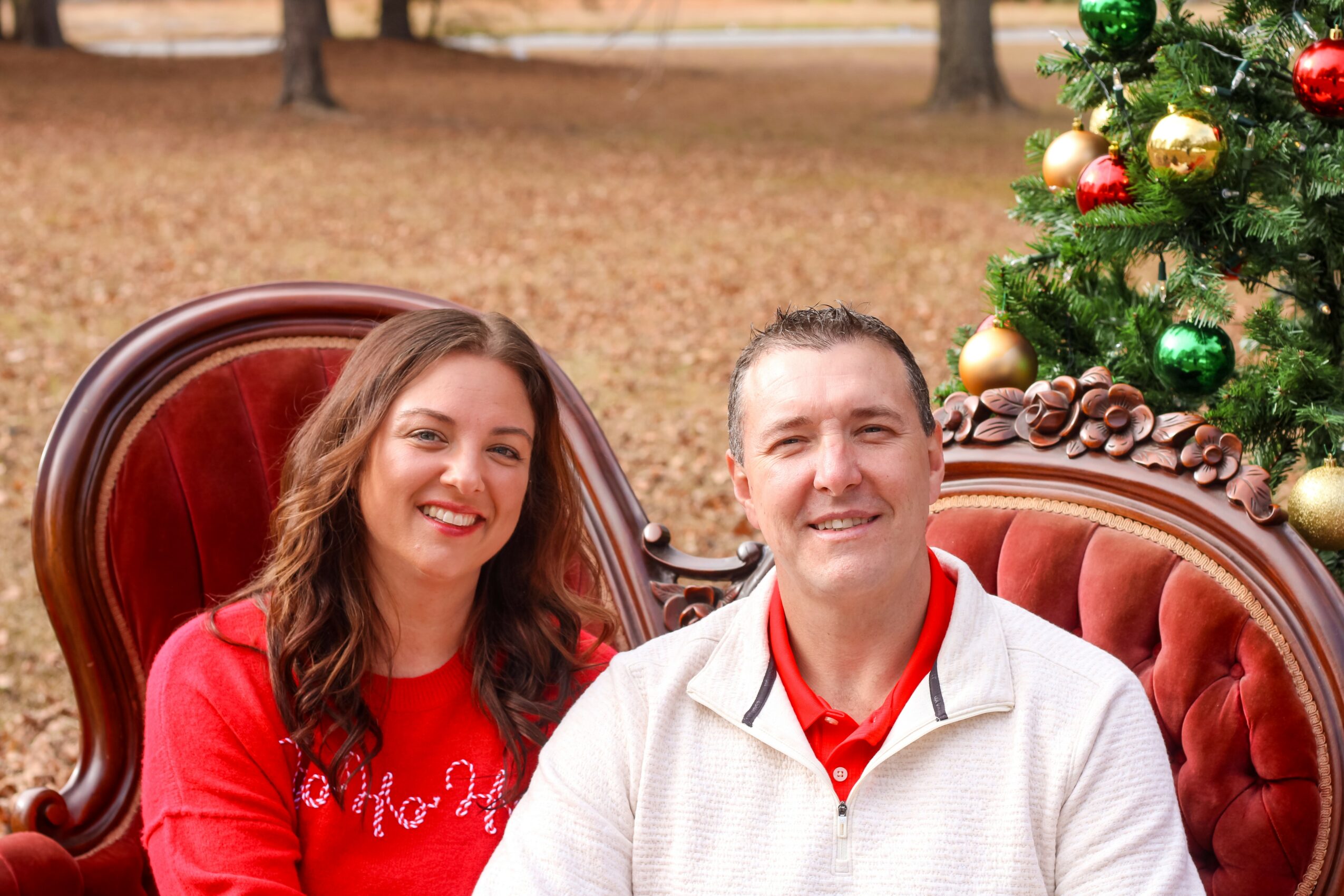 A smiling woman and man sit on a red velvet sofa outdoors with a decorated Christmas tree nearby.