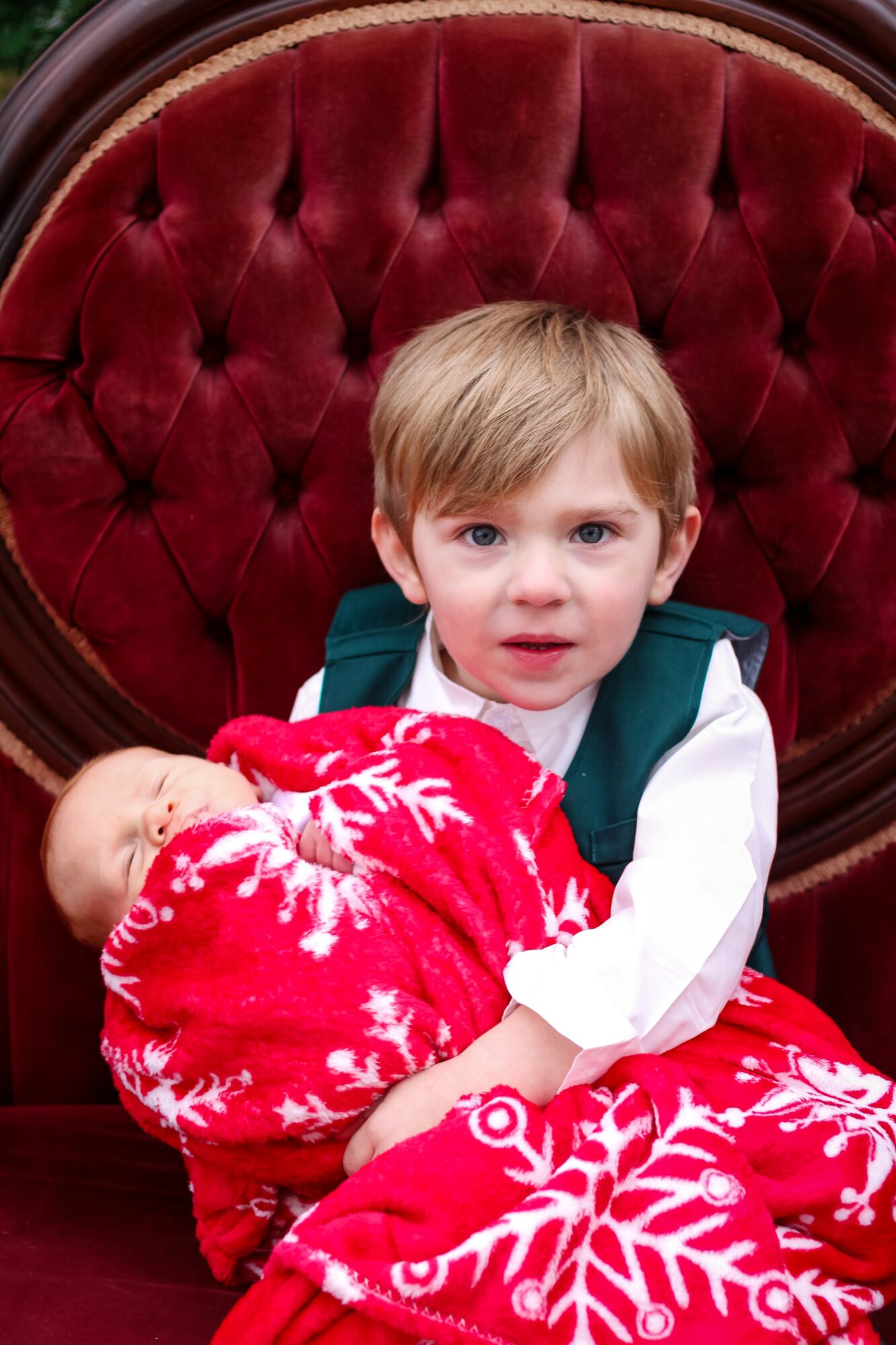 Young boy with light hair and blue eyes sitting on a red velvet chair, holding a red and white Christmas-themed blanket.