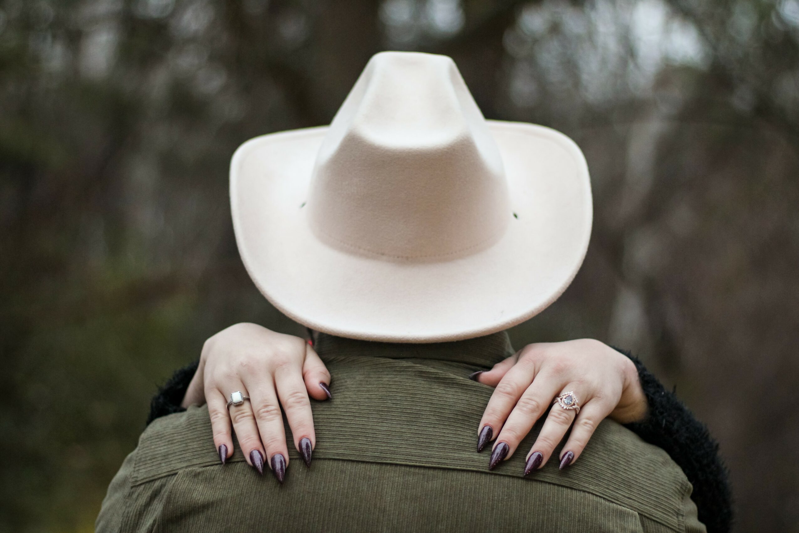 Person wearing a large white hat, holding onto the back of a jacket with hands visible, outdoors background.