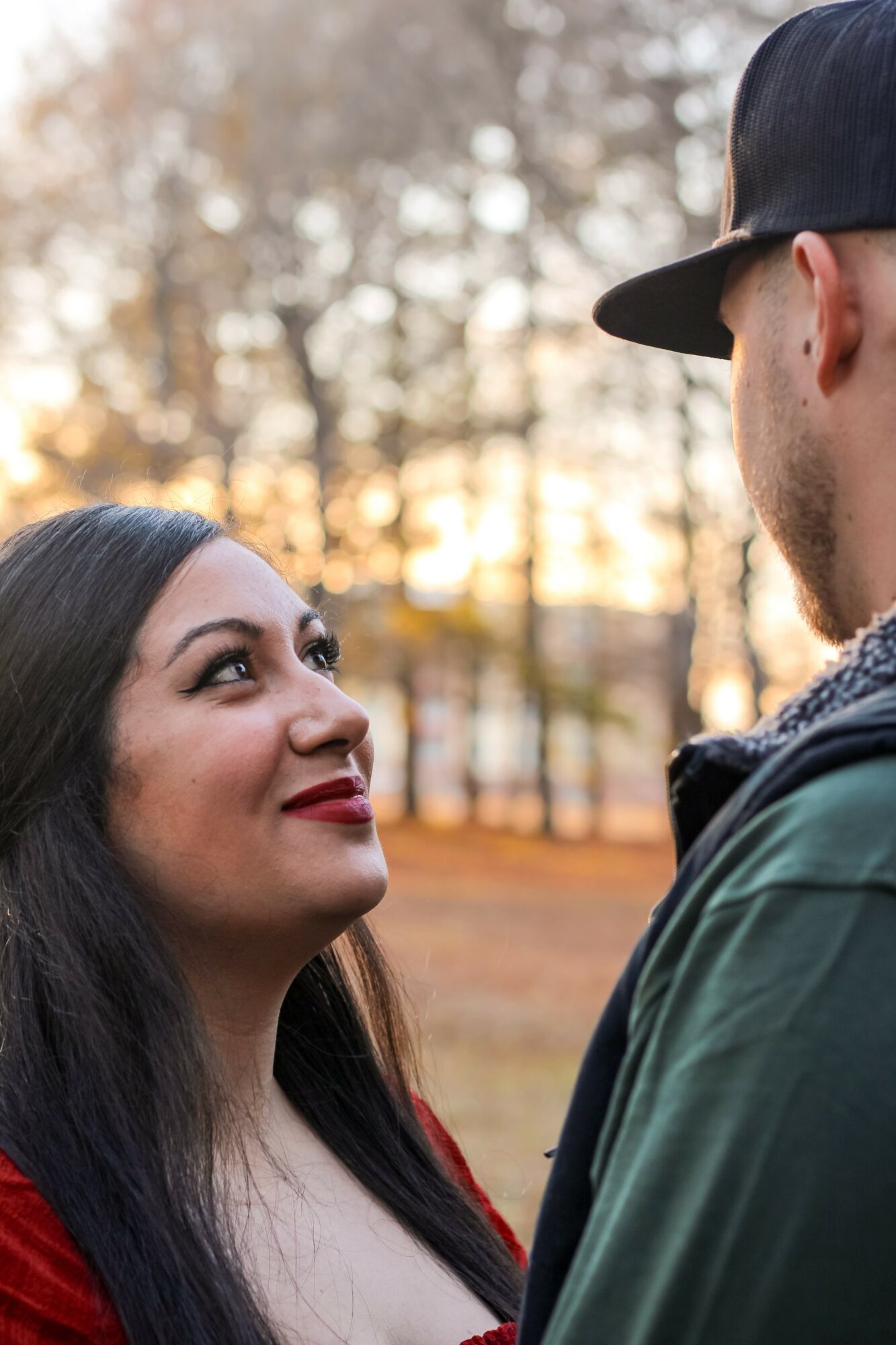 A woman and a man face each other outdoors with trees and sunlight in the background.