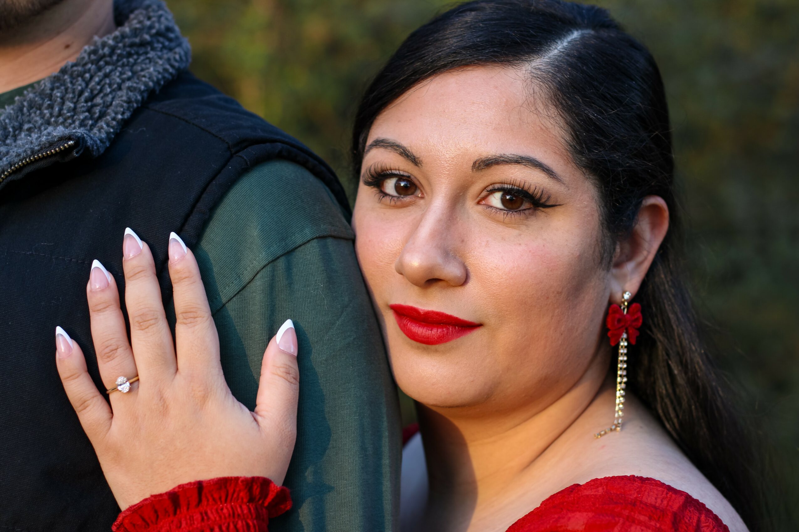 Woman with dark hair, red lipstick, and red earrings, smiling outdoors, with a hand on a person's shoulder.