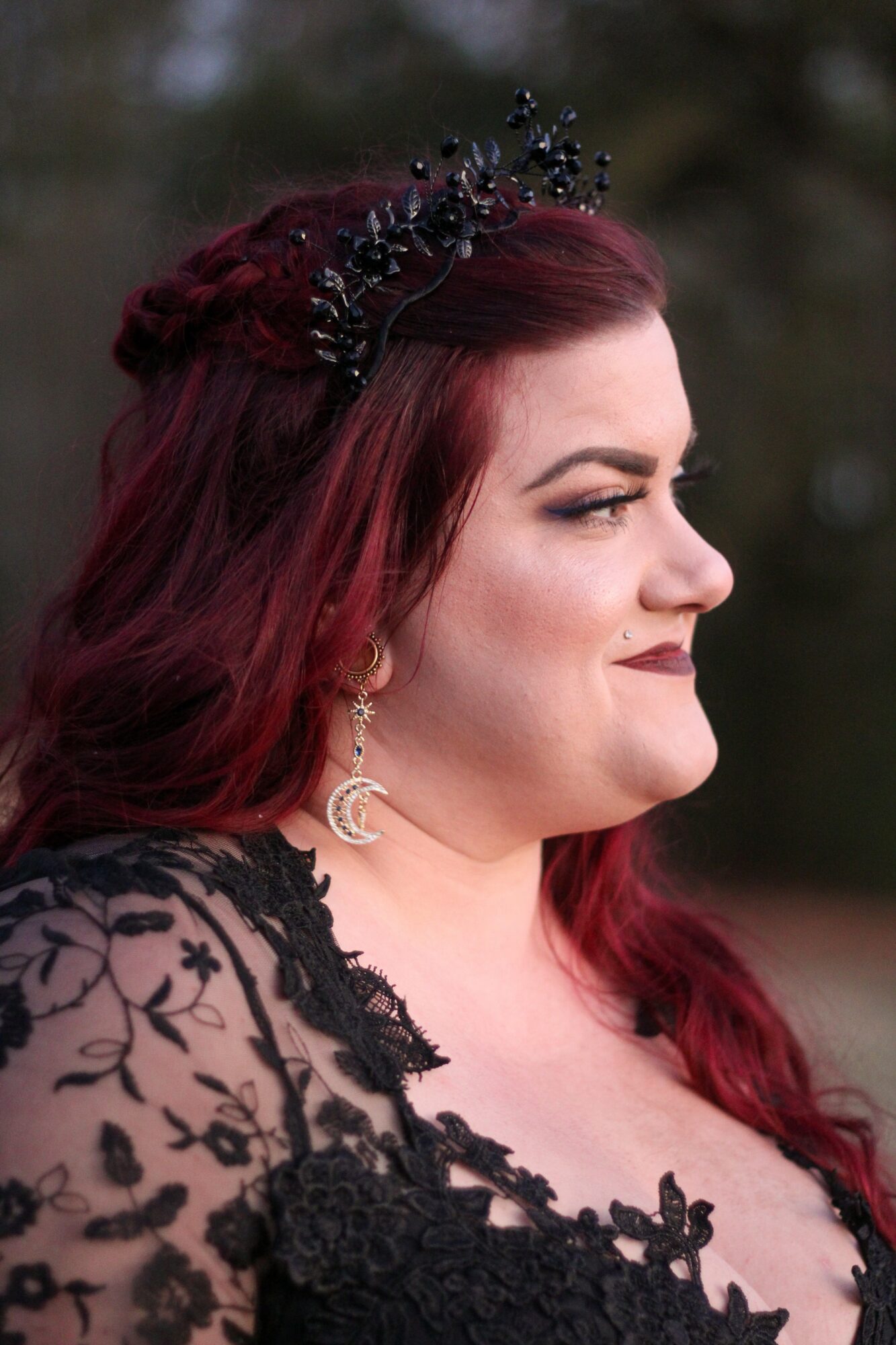 Side profile of a woman with long red hair, wearing a black lace dress and a decorative headpiece, outdoors.