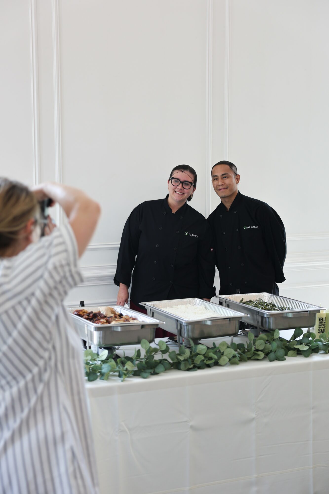 Two people in black uniforms stand behind a table with food trays, a person in striped shirt takes a photo.