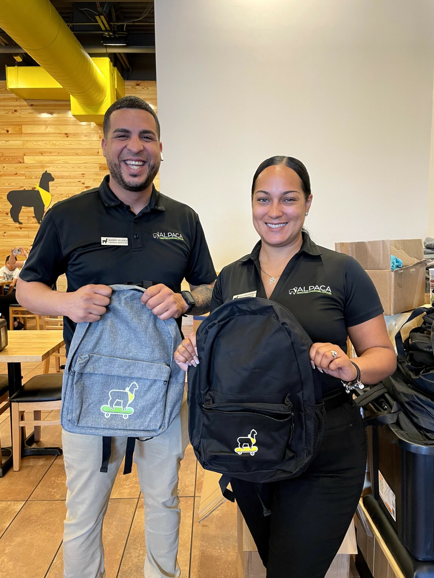Two smiling employees holding backpacks inside a store, one with a gray backpack and the other with a black backpack.