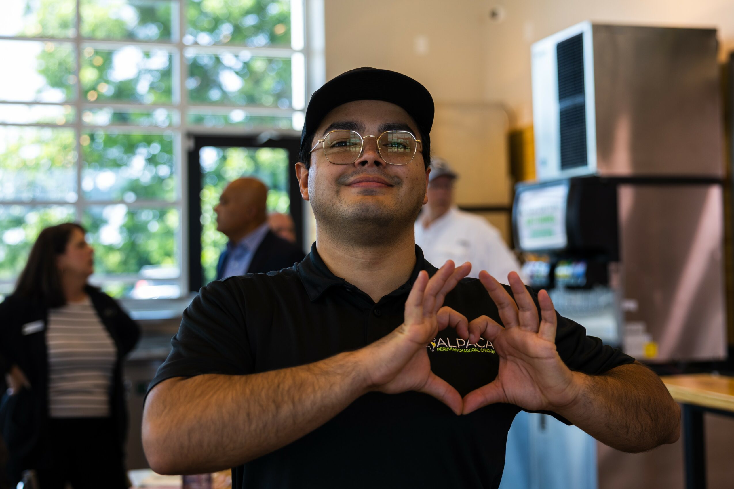Person smiling and making a heart shape with hands in a cafe or restaurant, with people and windows in background.
