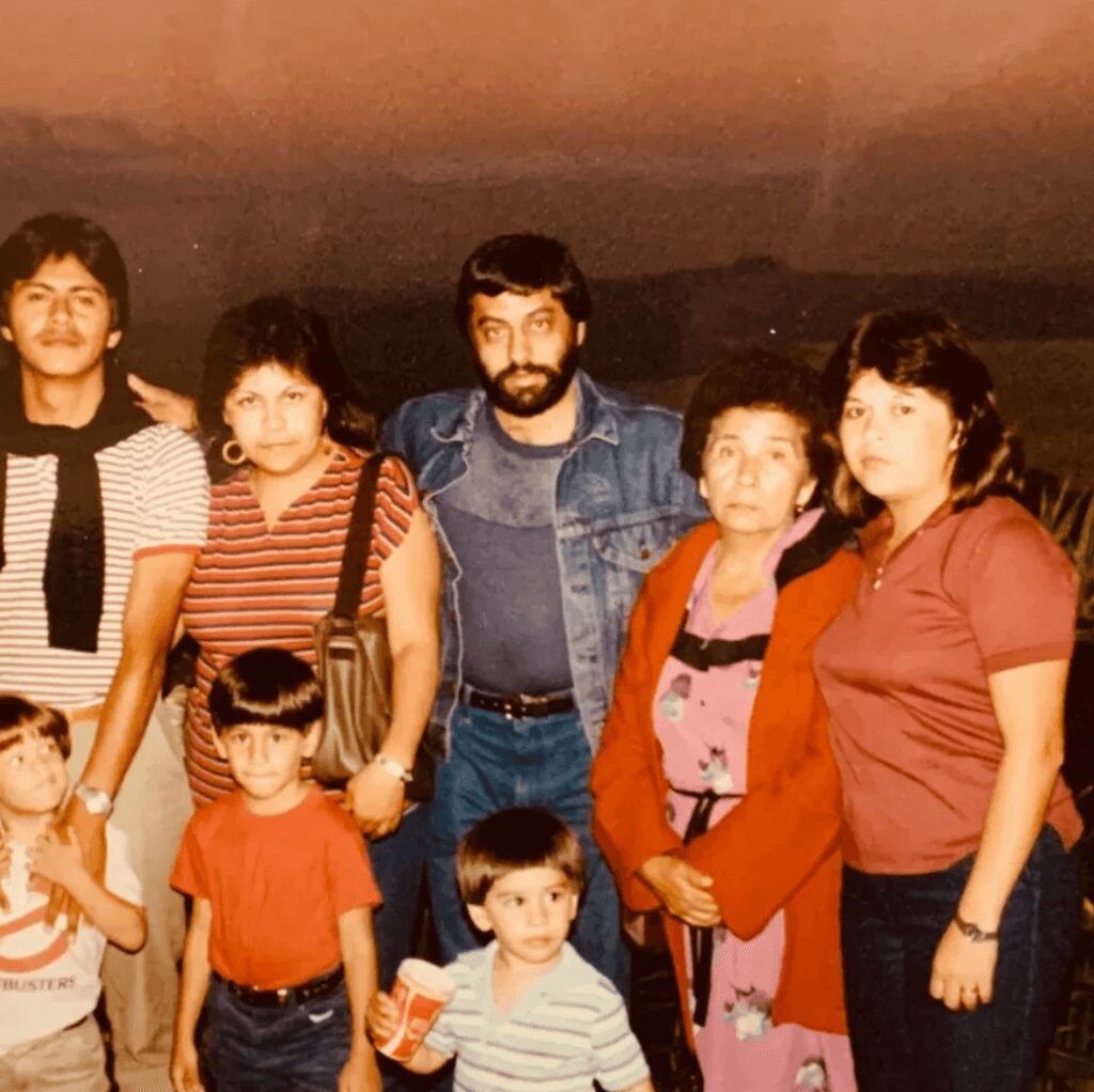 Group of seven people, including children and adults, standing together indoors. Some are smiling, wearing casual clothing.