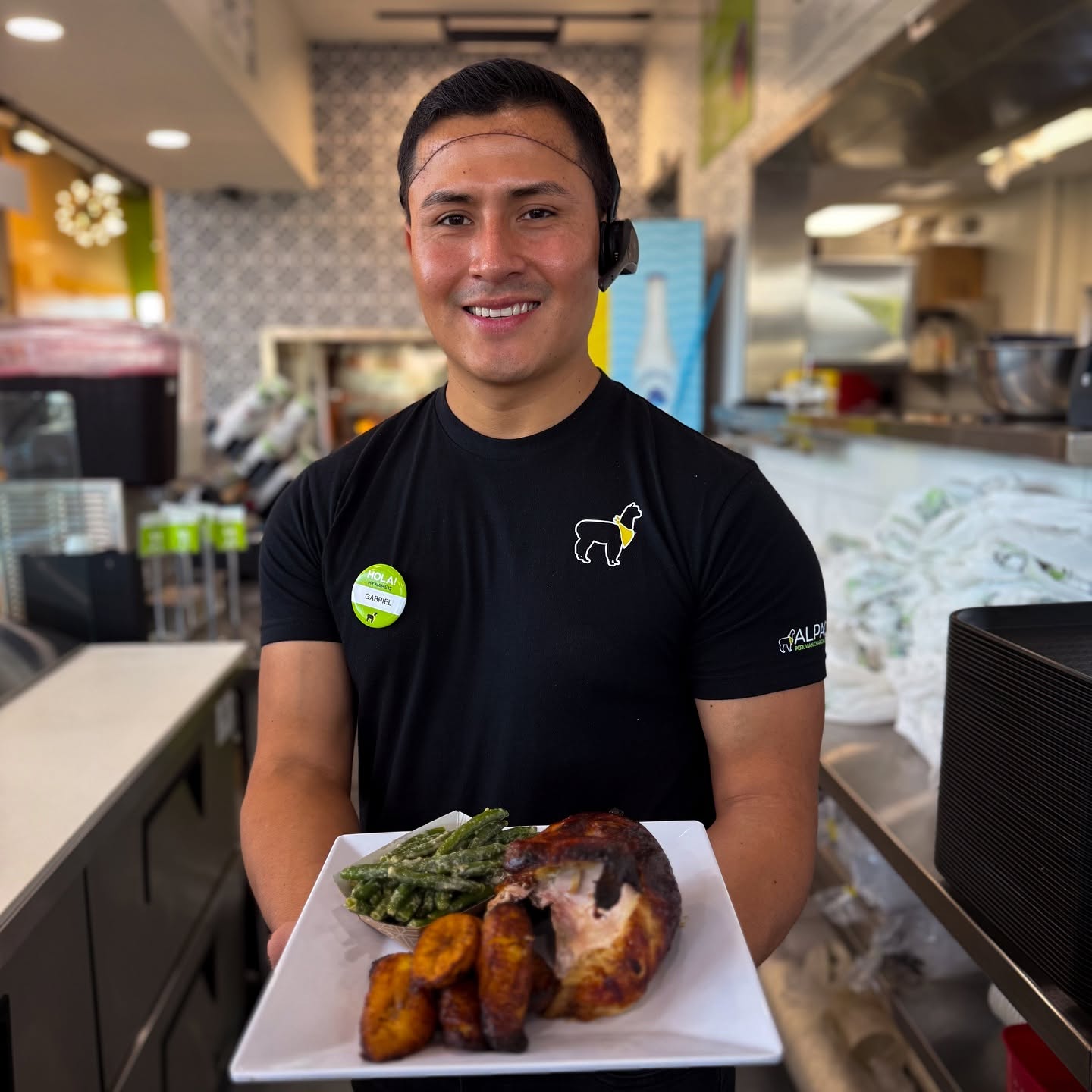 A smiling person in a black shirt holding a plate with meat, vegetables, and fried items in a restaurant.