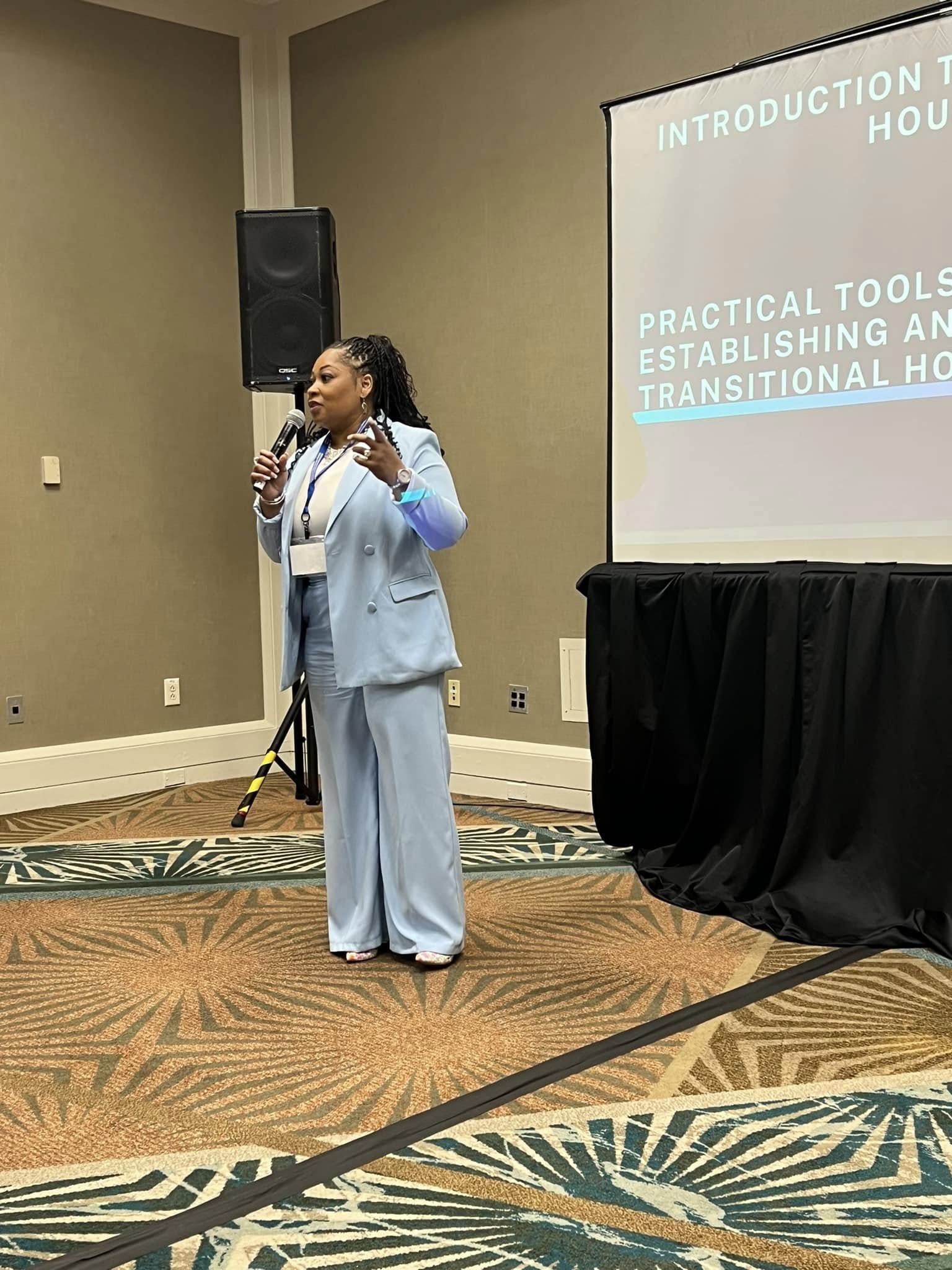 Woman in light blue suit speaking into microphone during presentation in conference room.