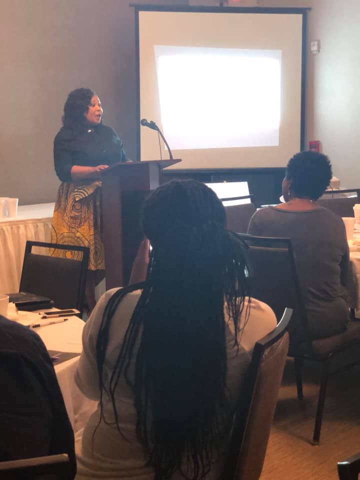 Woman standing at podium giving presentation to seated audience in a conference room.