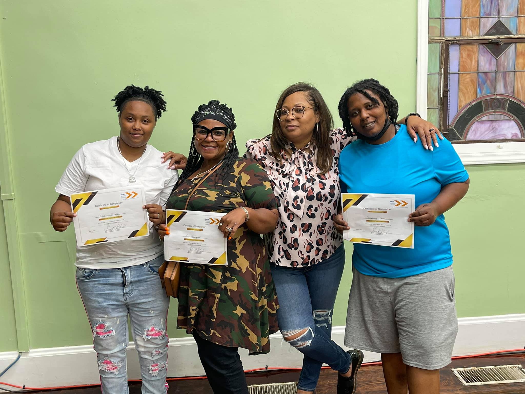 Four women standing indoors, holding certificates, smiling, with a green wall and a window in the background.