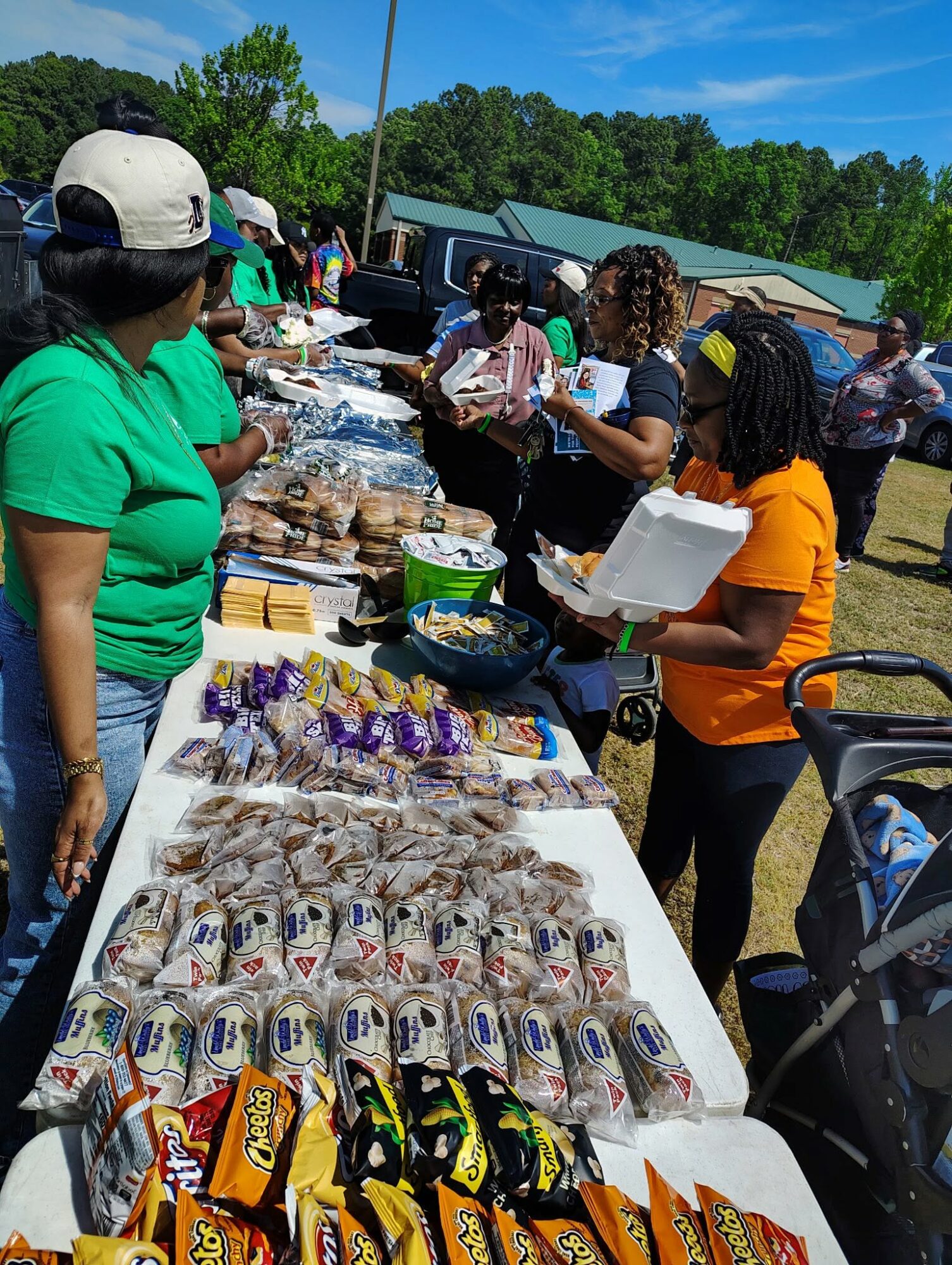 People gather around a table with baked goods and snacks outdoors on a sunny day.