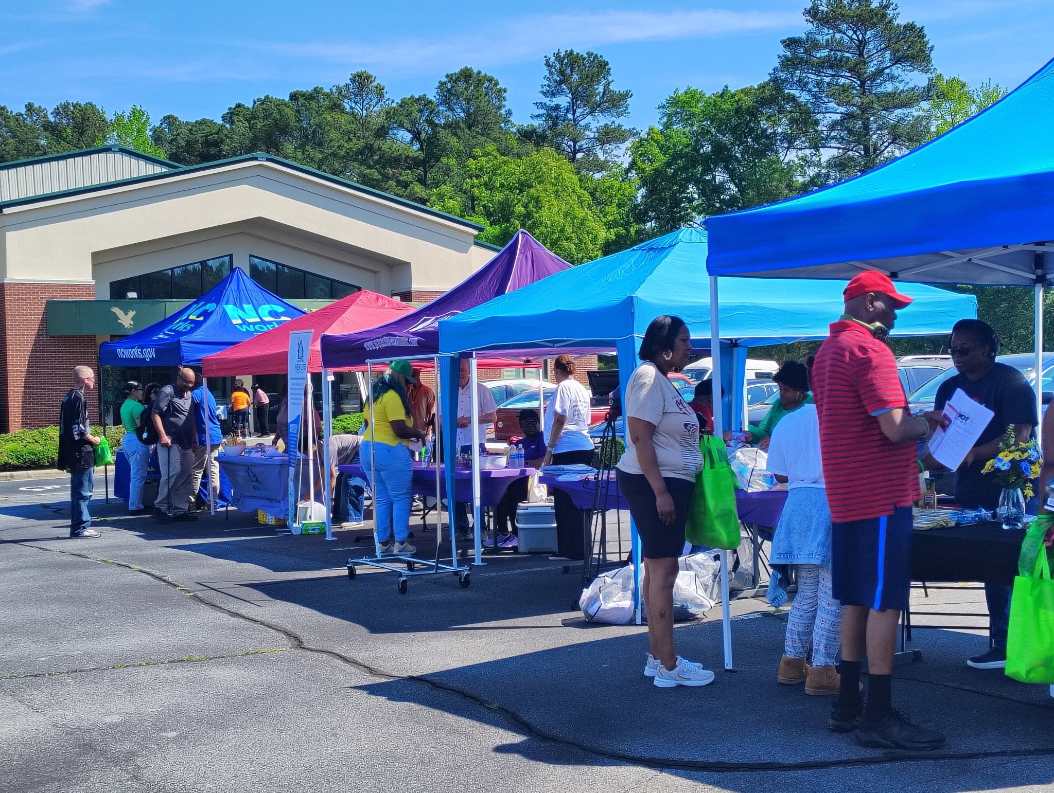 People browsing outdoor market stalls with colorful tents on a sunny day, trees and a building in the background.