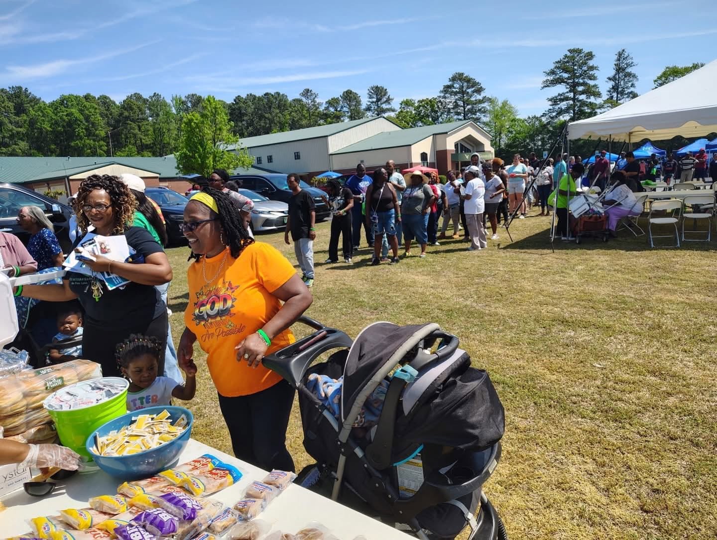 People gather outdoors near tables and tents, with a building and trees in the background, on a sunny day.