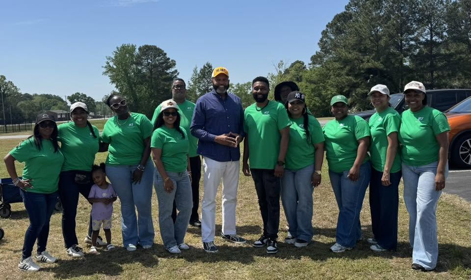 Group of people standing outdoors on grass, some wearing green shirts, with trees and a blue sky in background.