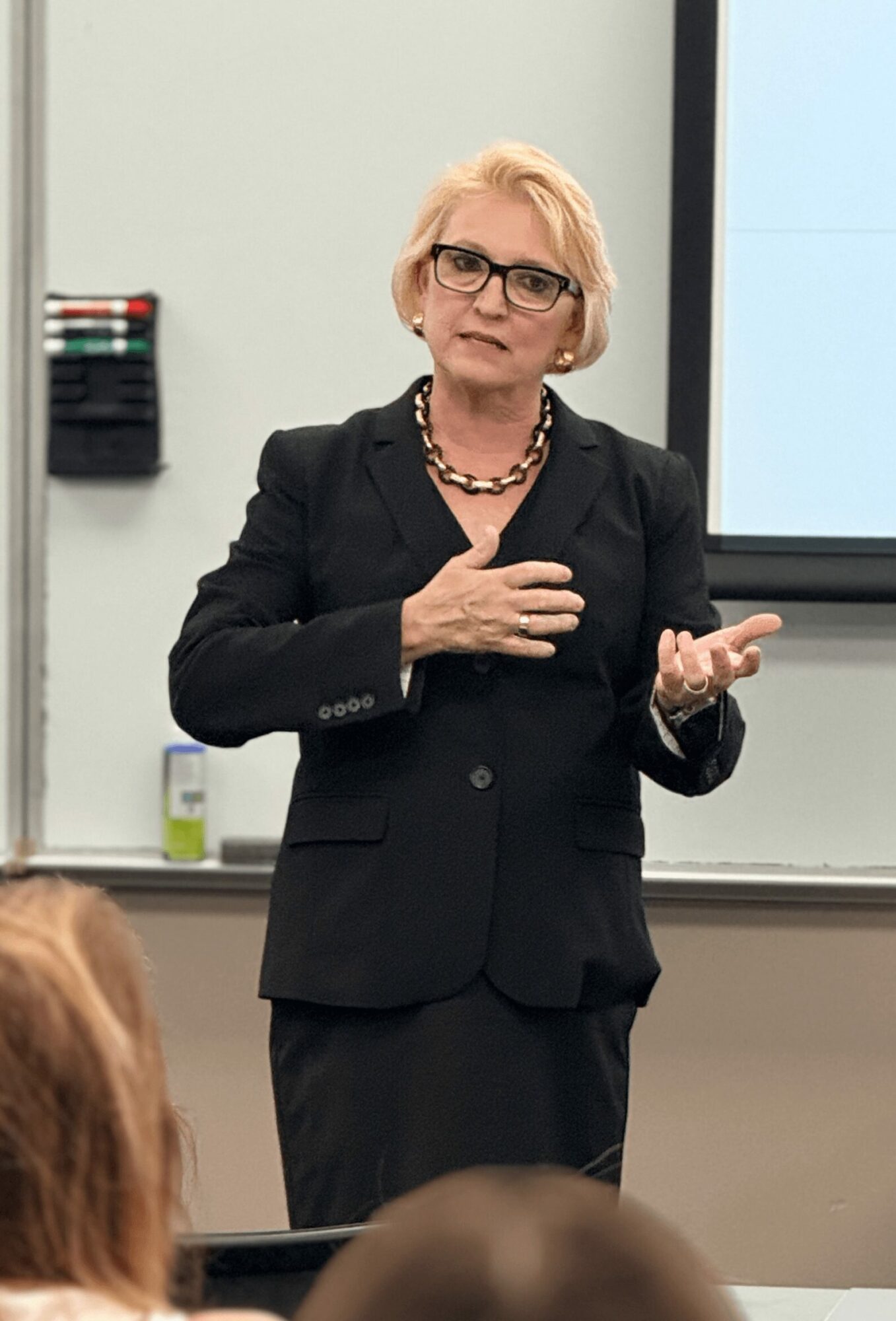 Woman with glasses and blonde hair speaking in a classroom, gesturing with her right hand, wearing a black suit and necklace.