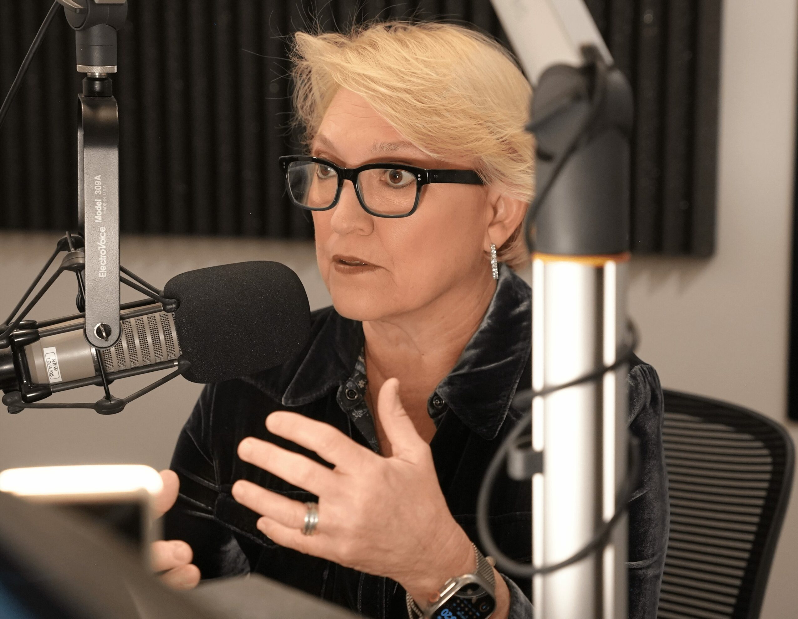 Woman with short blonde hair, glasses, and earrings speaking into a microphone in a recording studio.