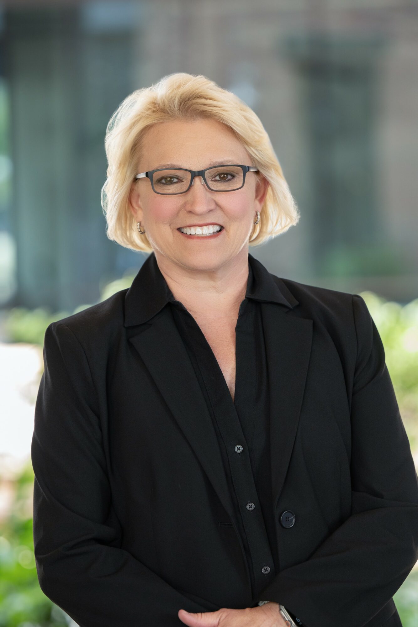 Woman with blonde hair, glasses, wearing a black blazer, smiling outdoors with a blurred background.