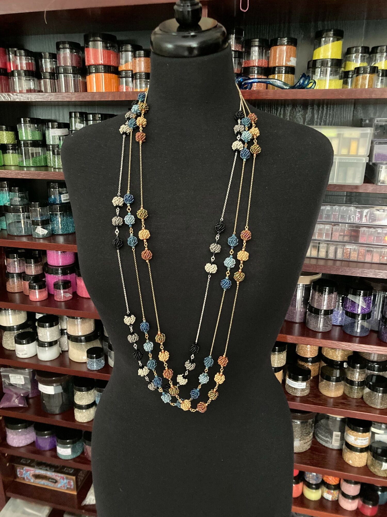 Necklace with multiple strands of beads displayed on a black mannequin, background shows shelves of colorful thread spools.