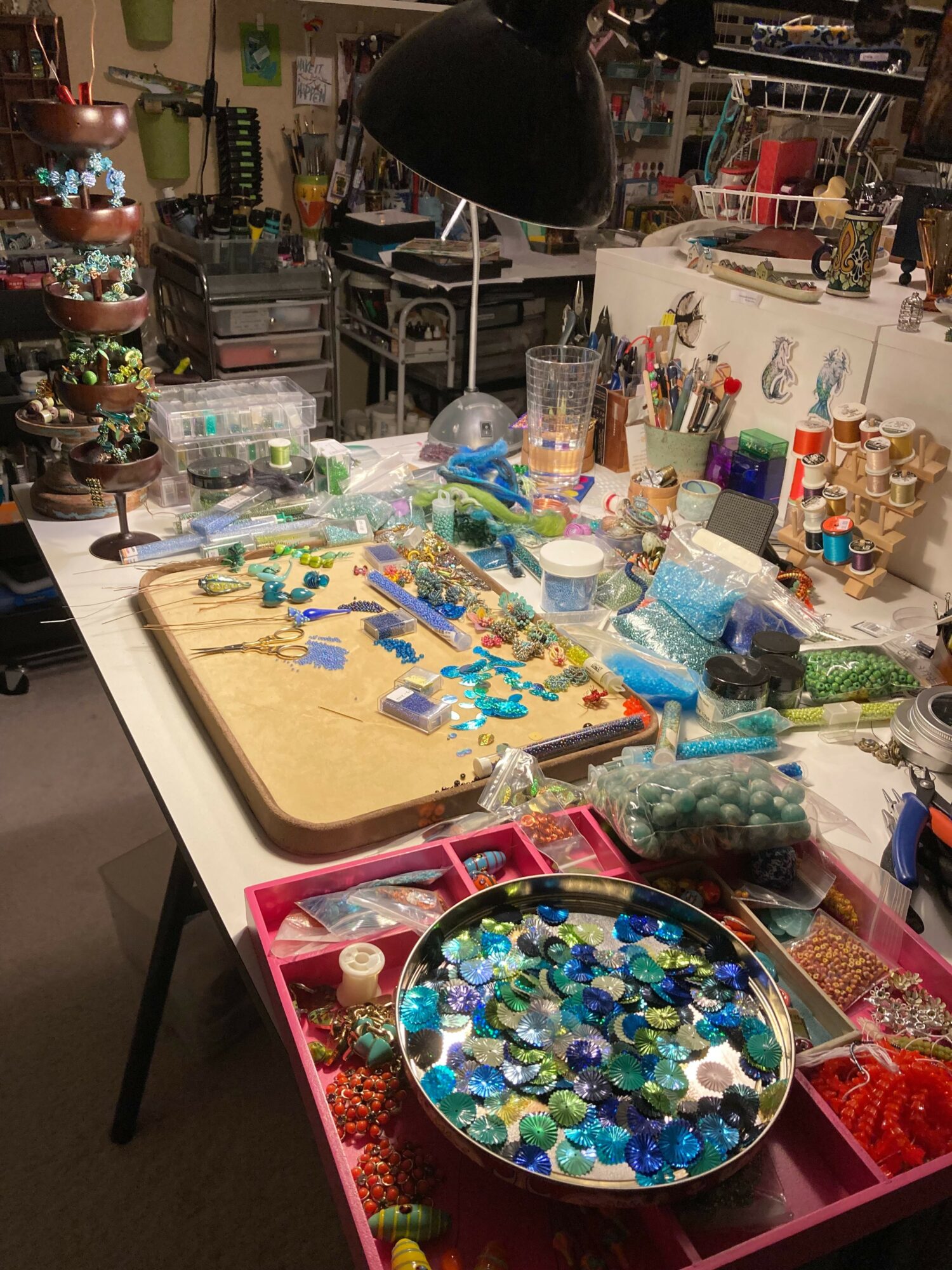 Workstation with beads, tools, and supplies for jewelry making, illuminated by a hanging lamp, with organized storage in the background.