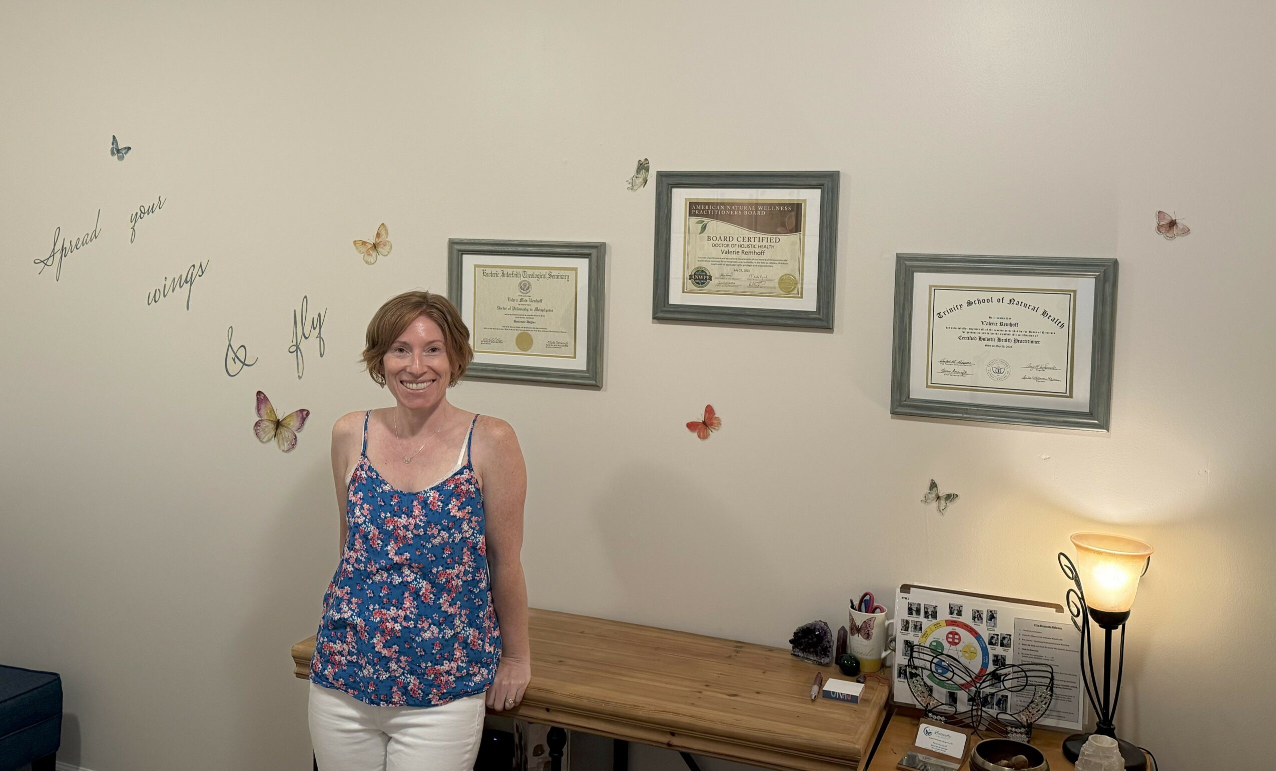 Woman standing in front of a wall with framed certificates and butterfly decorations, smiling, with a desk and lamp nearby.