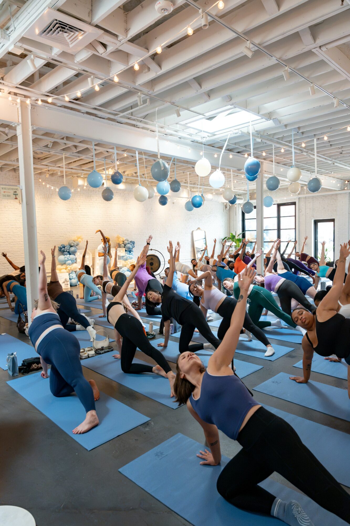 Group of people practicing yoga in a spacious studio with hanging blue and white balls, large windows, and high ceiling.