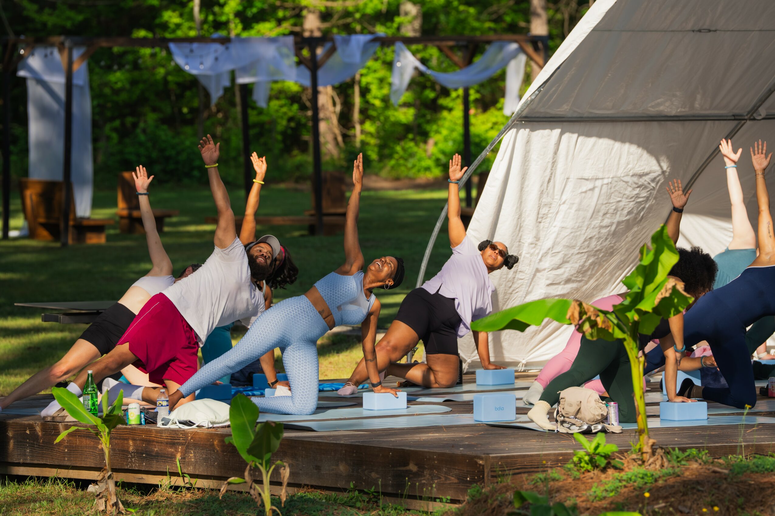 Group of people practicing yoga outdoors on a wooden platform, with arms raised and bodies in various poses, surrounded by greenery.