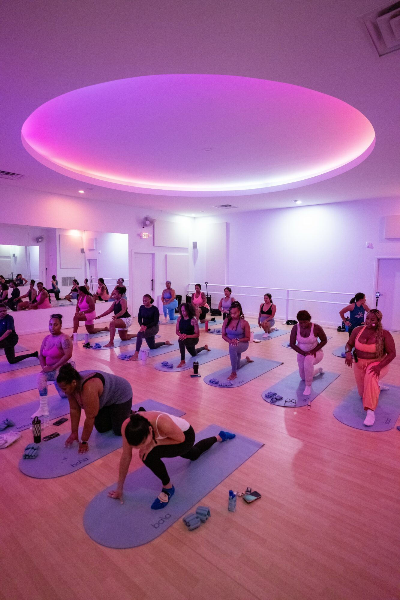 Group of people participating in a yoga class in a spacious room with purple lighting, mats, and a circular ceiling light.