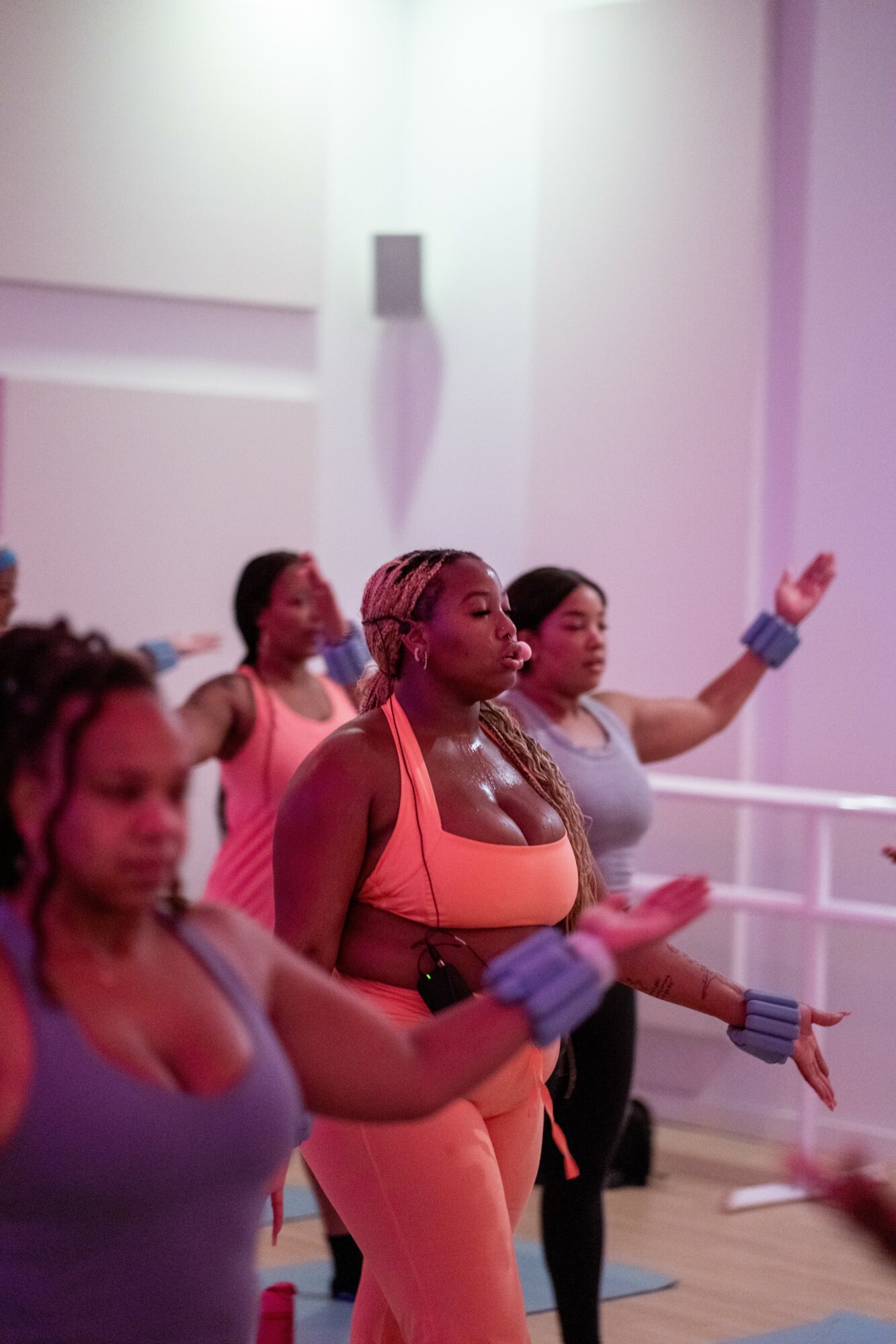 Group of women practicing yoga or meditation in a studio with soft lighting, some with eyes closed and hands raised.