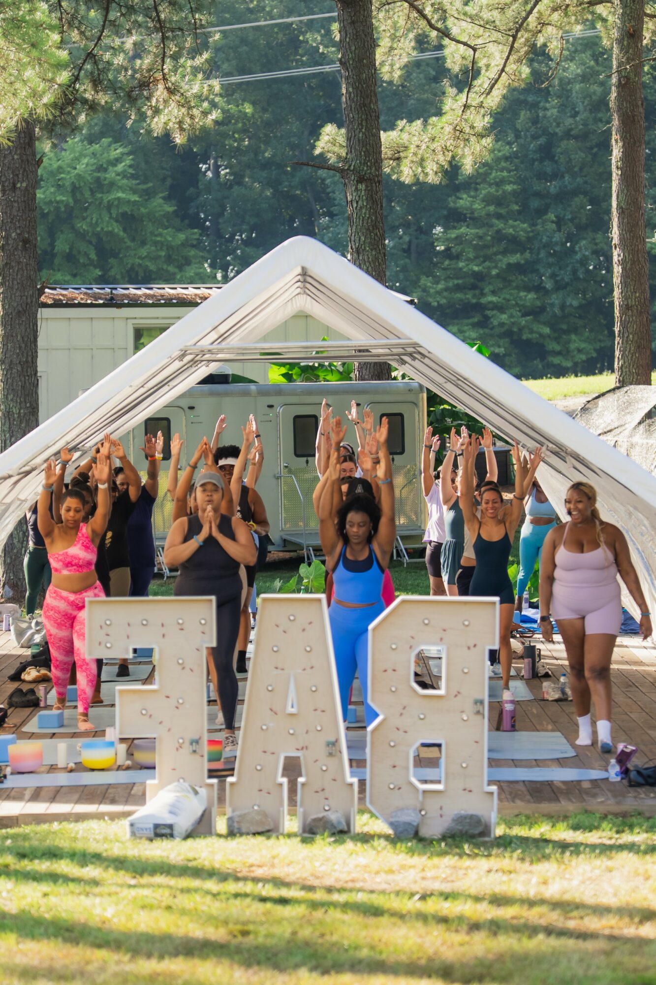 Group of people practicing yoga outdoors under a white tent, with large letters spelling 'PEACE' in front, trees in background.