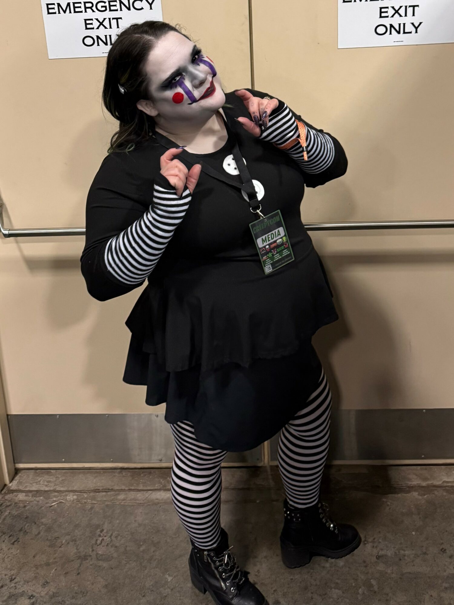Person with face paint, striped clothing, and a badge, standing in front of emergency exit signs, making fists.