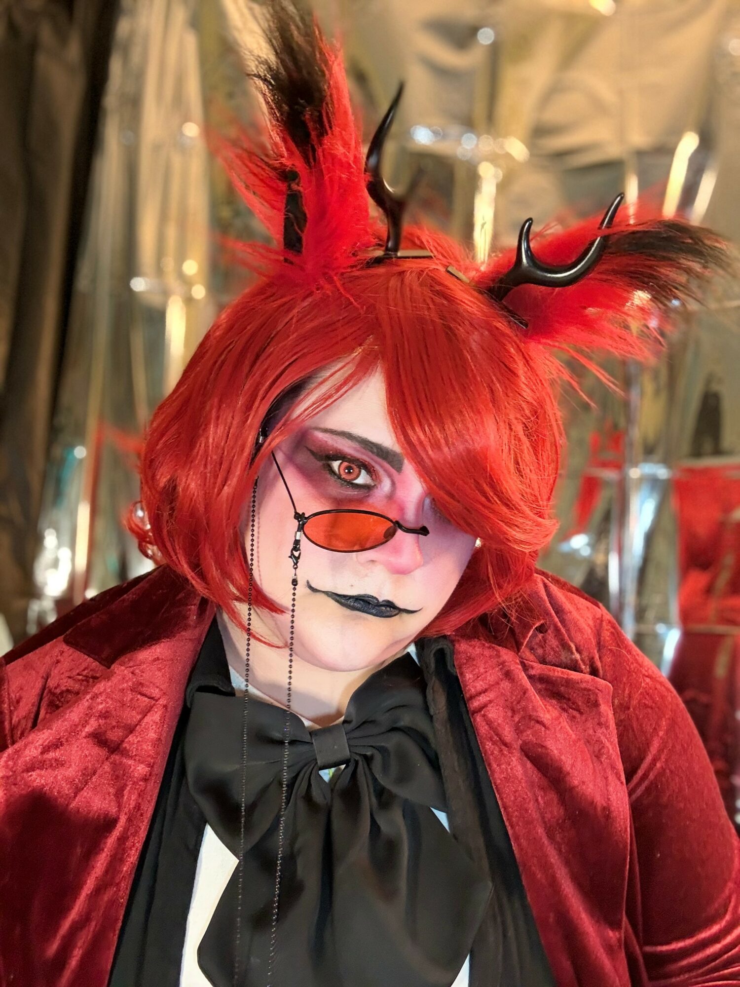 Person with bright red hair, fox-like ears, and black horns, wearing sunglasses, black lipstick, and a velvet red jacket.