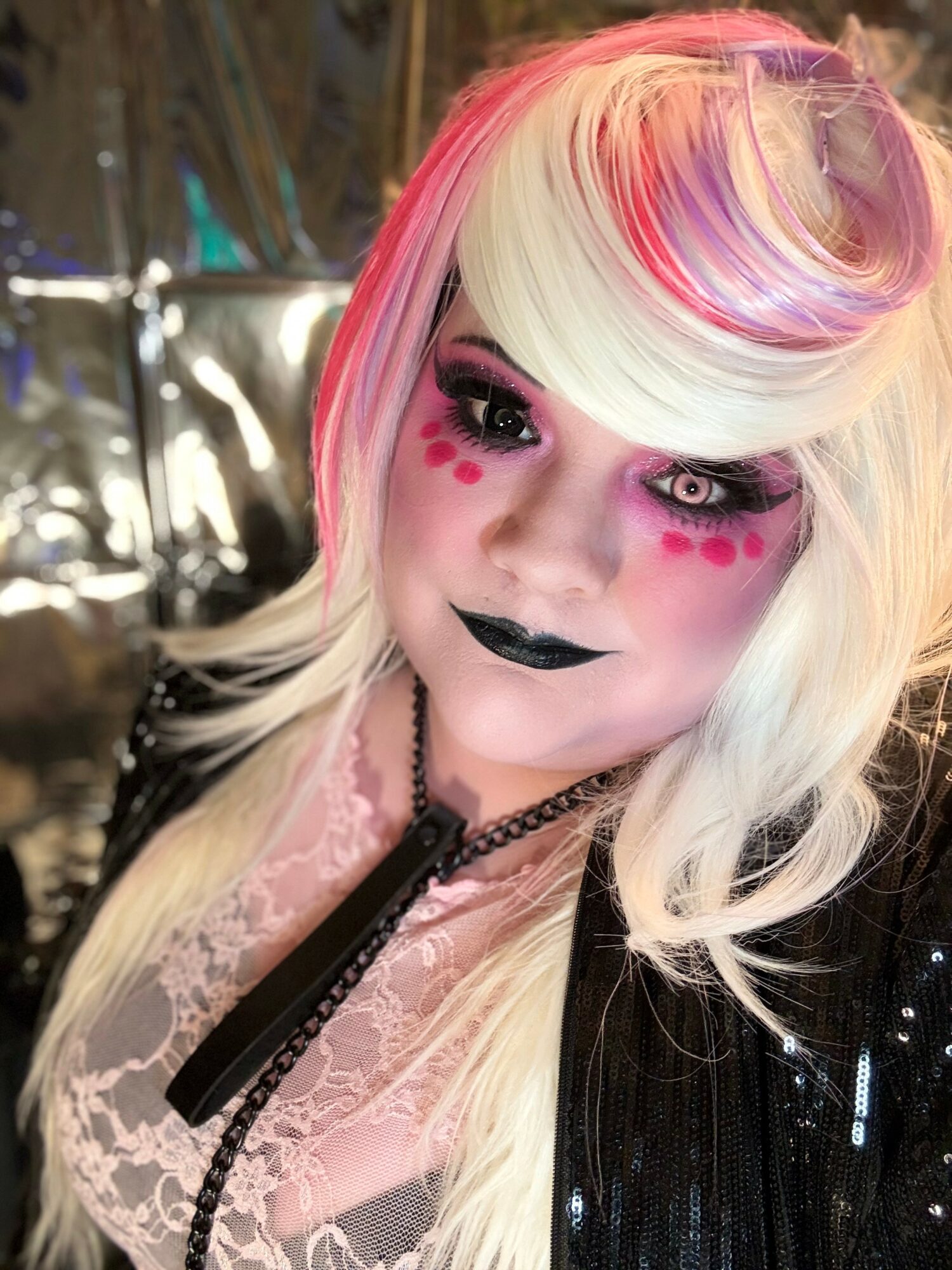 Person with colorful hair, makeup, and black lipstick, wearing a lace top and black jacket, in a dimly lit setting.