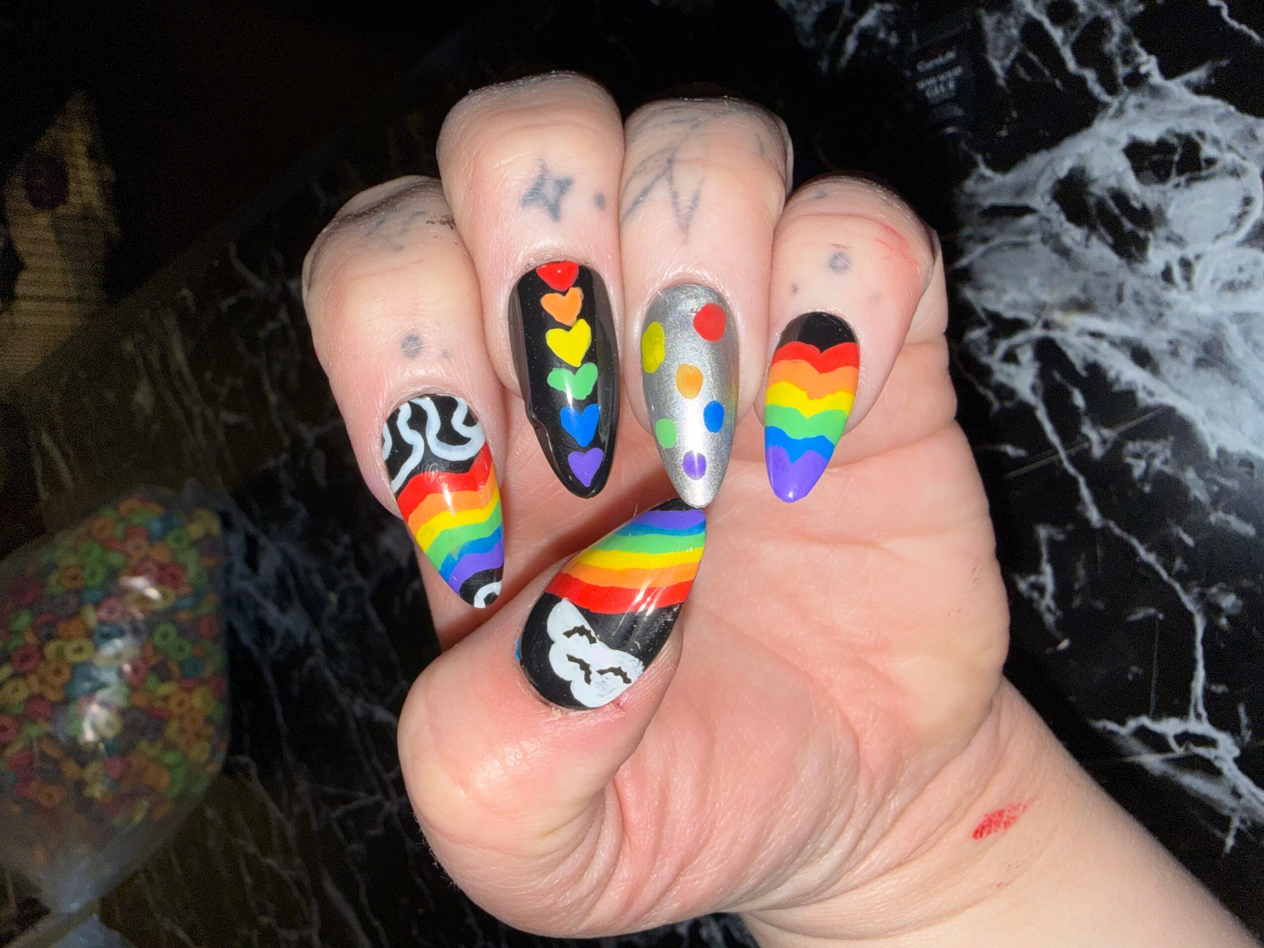 Hand with rainbow-themed nail art, including hearts, stars, and rainbow stripes, against a dark background.