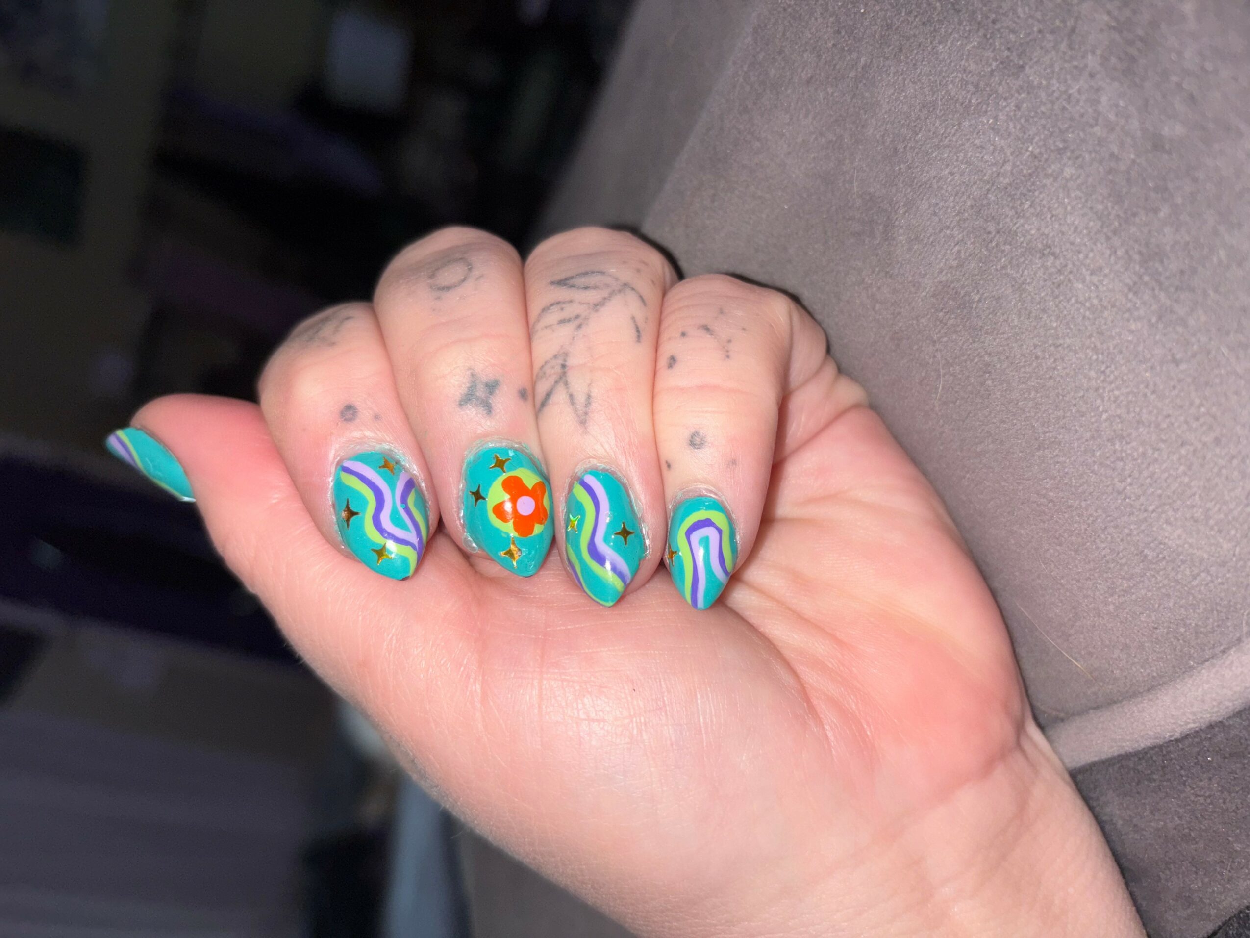 Hand with colorful, patterned nail art on fingernails, tattoos on fingers, resting on a soft surface.