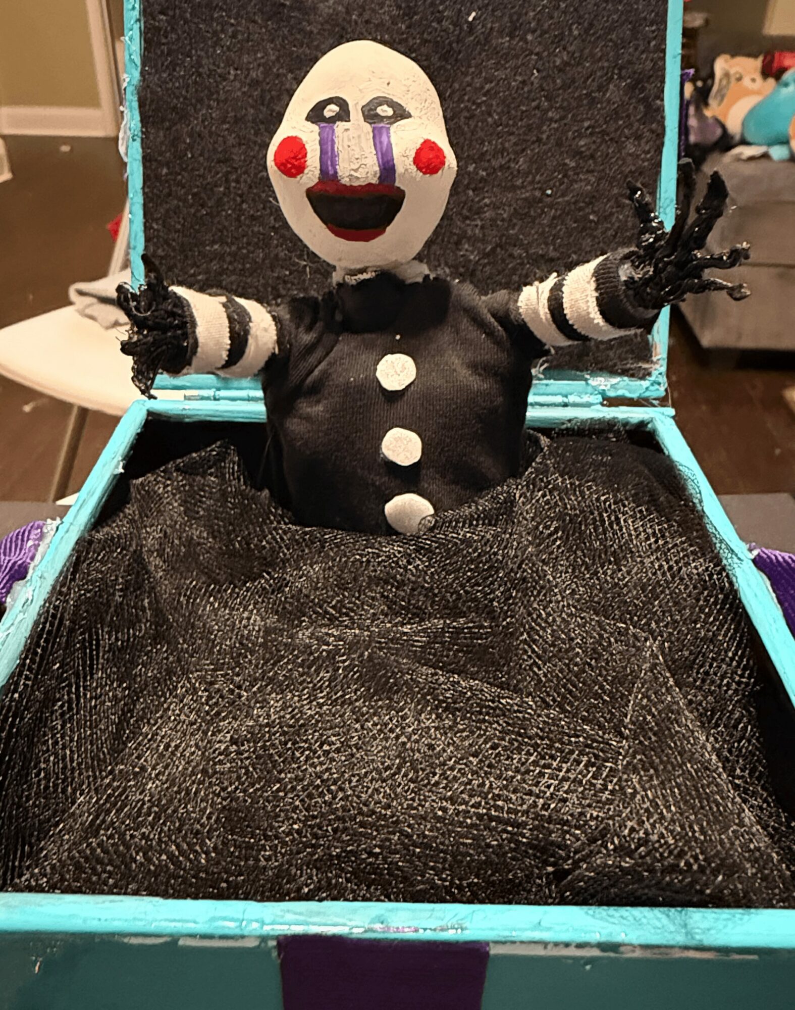 Clown puppet with a white face, red cheeks, and a big smile, dressed in black, inside a box with a black fabric lining.
